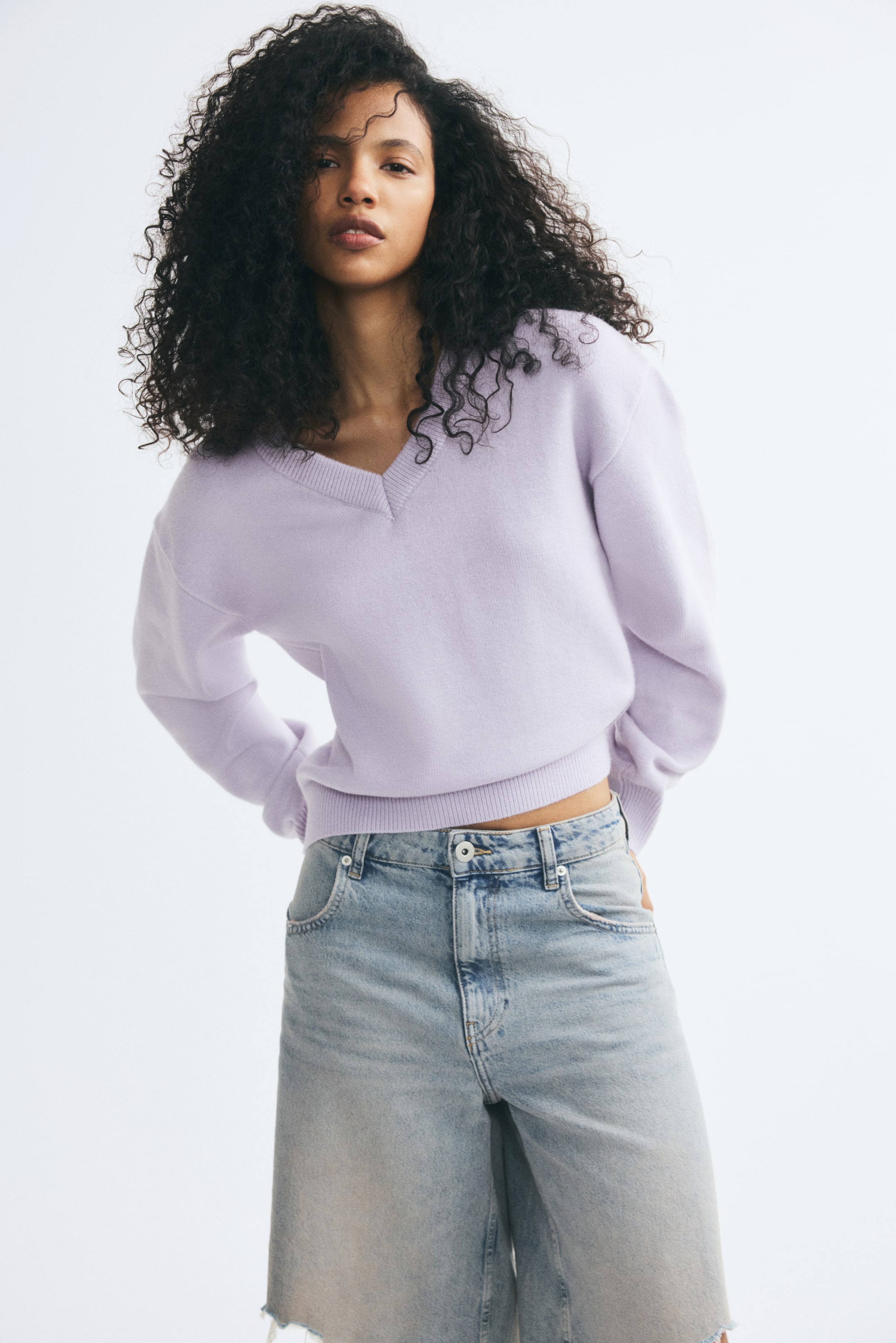Visualizza l'immagine ingrandita: Un maglione in maglia lilla chiaro, caratterizzato da un collo a V a coste, spalle scese e fondo a coste, indossato dalla modella. Il maglione è abbinato a jeans in denim dal lavaggio chiaro.