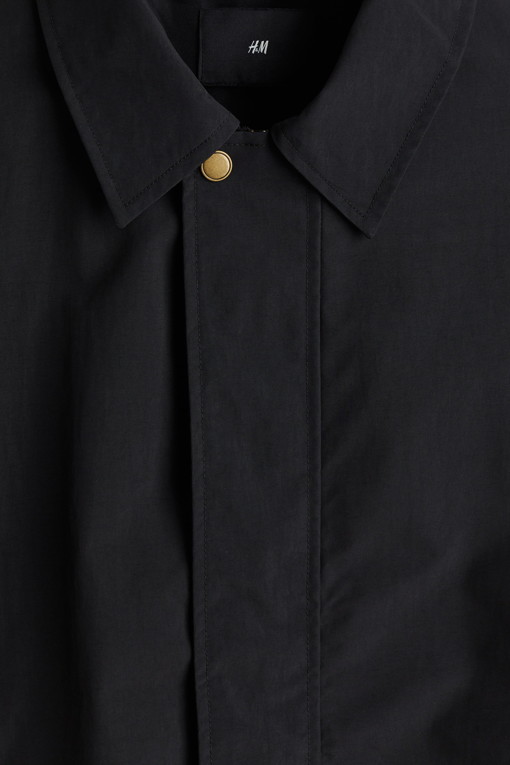 拡大画像を表示: シャープなナイロン製の黒いジャケットが前面から紹介され、尖った襟が特徴です。中央には風よけのフラップがあり、目に見える真鍮色のプレススタッドで固定されています。