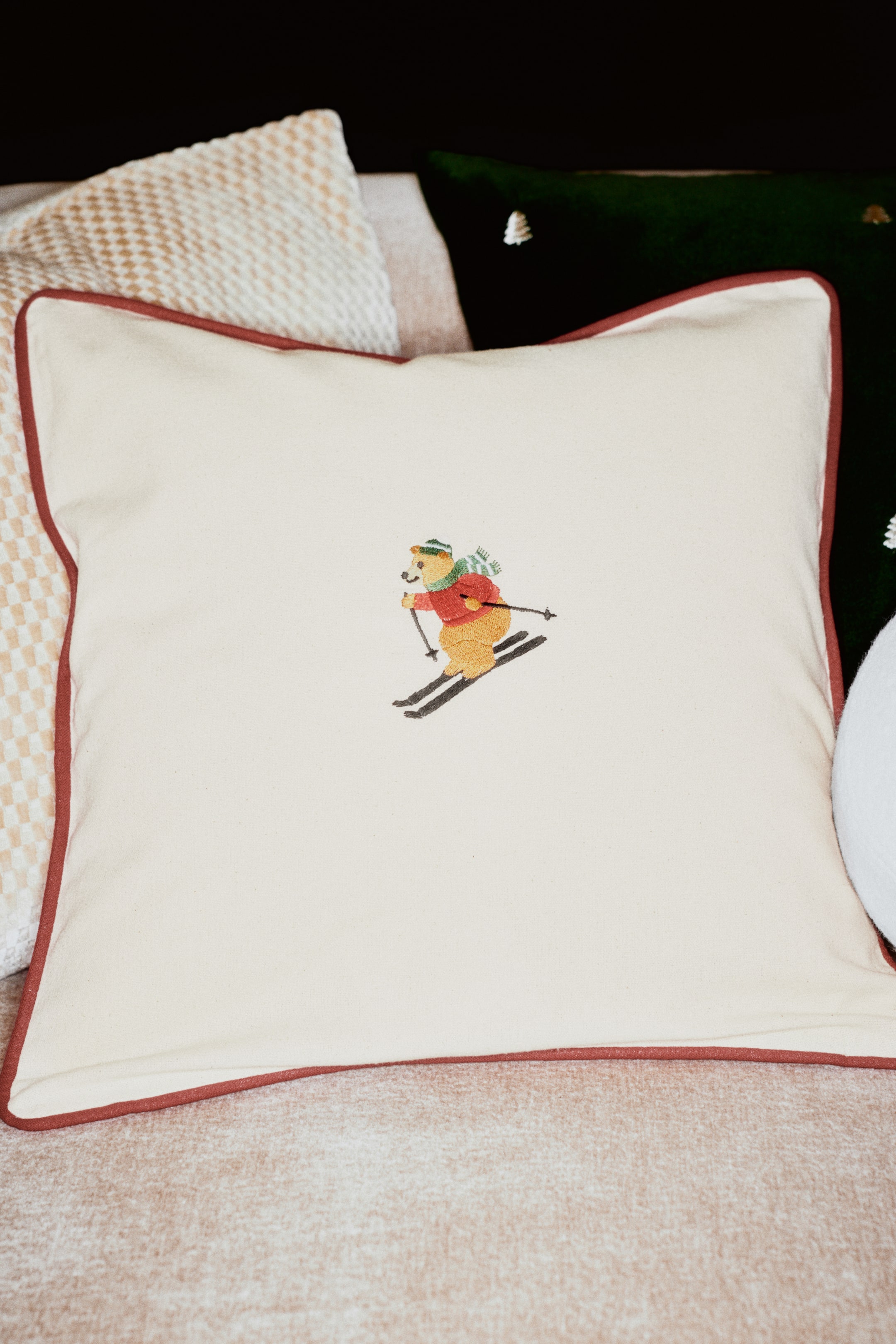 View larger image: A cream-coloured cotton canvas cushion cover is shown from the front, featuring a small embroidered detail of a skiing bear in the centre. The square cover is finished with contrasting reddish-brown piping along the edges.