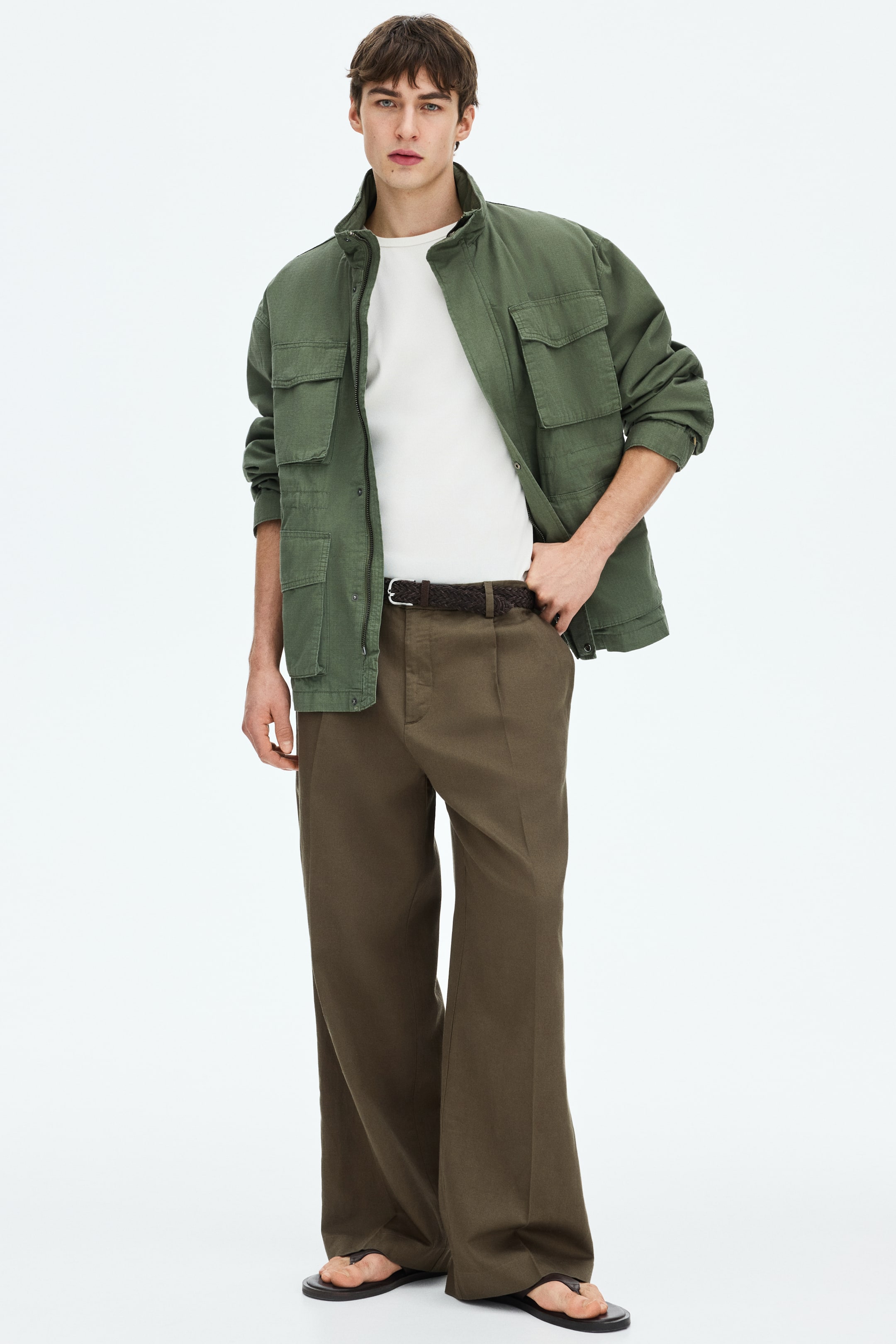 View larger image: An olive green utility jacket, worn open over a white crew-neck t-shirt, is showcased from a front-facing angle. The jacket features a standing collar, a front zipper, and four flap pockets, and is paired with brown wide-leg pleated trousers, a dark brown braided belt, and dark flip-flops.