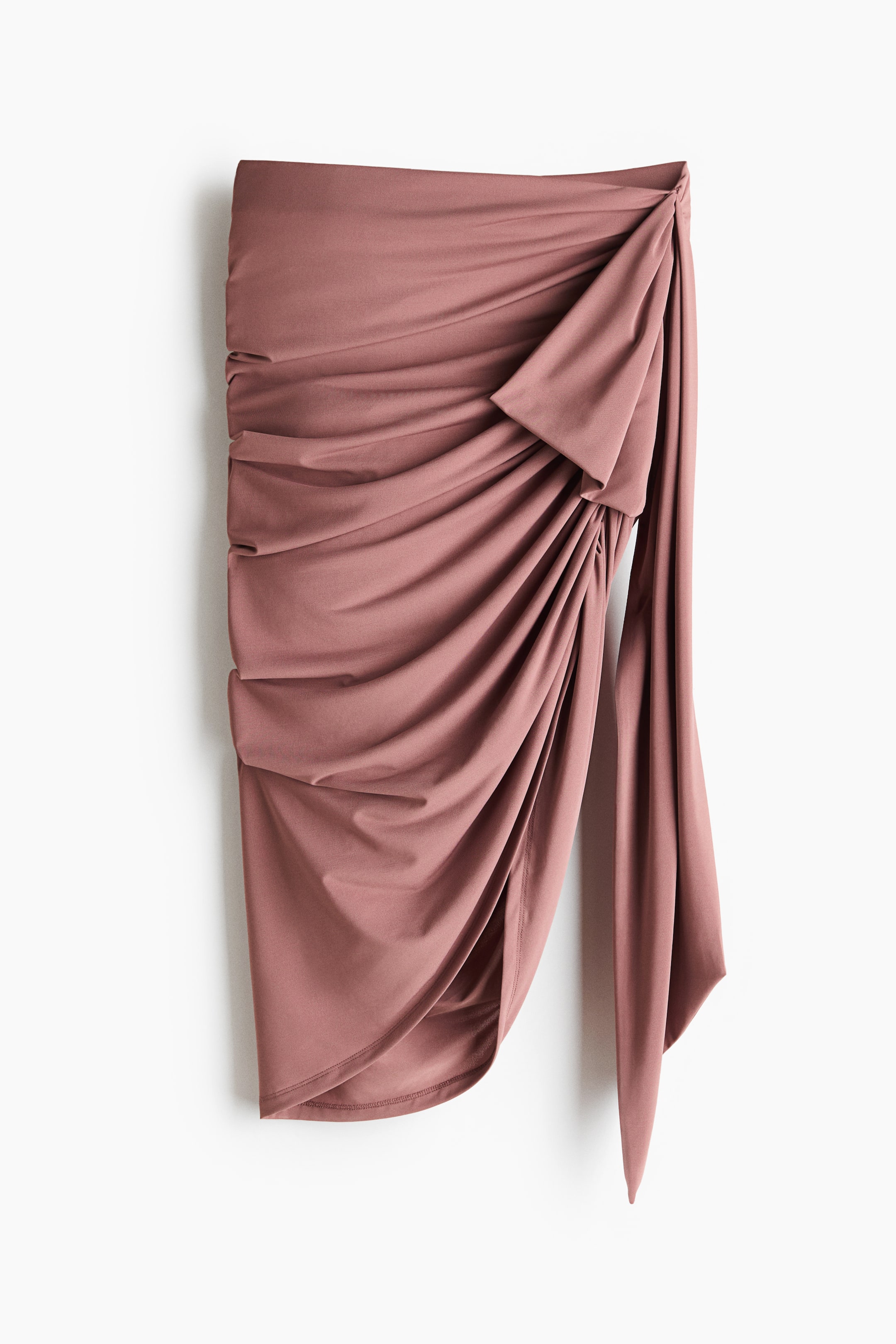 View larger image: A fitted skirt in jersey fabric is shown from the front, featuring a high waist. Decorative ties create a gathered, draped effect on one side, forming a rounded, asymmetric hem.
