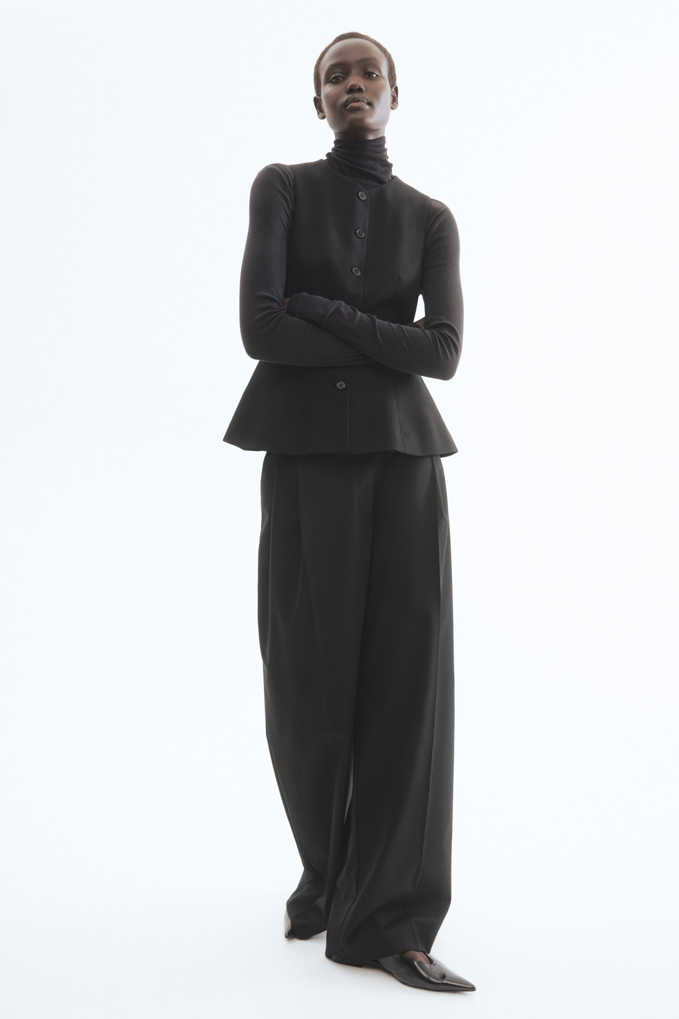 View larger image: The black ensemble showcases a high-neck, long-sleeve top layered beneath a peplum jacket with front buttons, complemented by wide-leg trousers and pointed-toe flats.