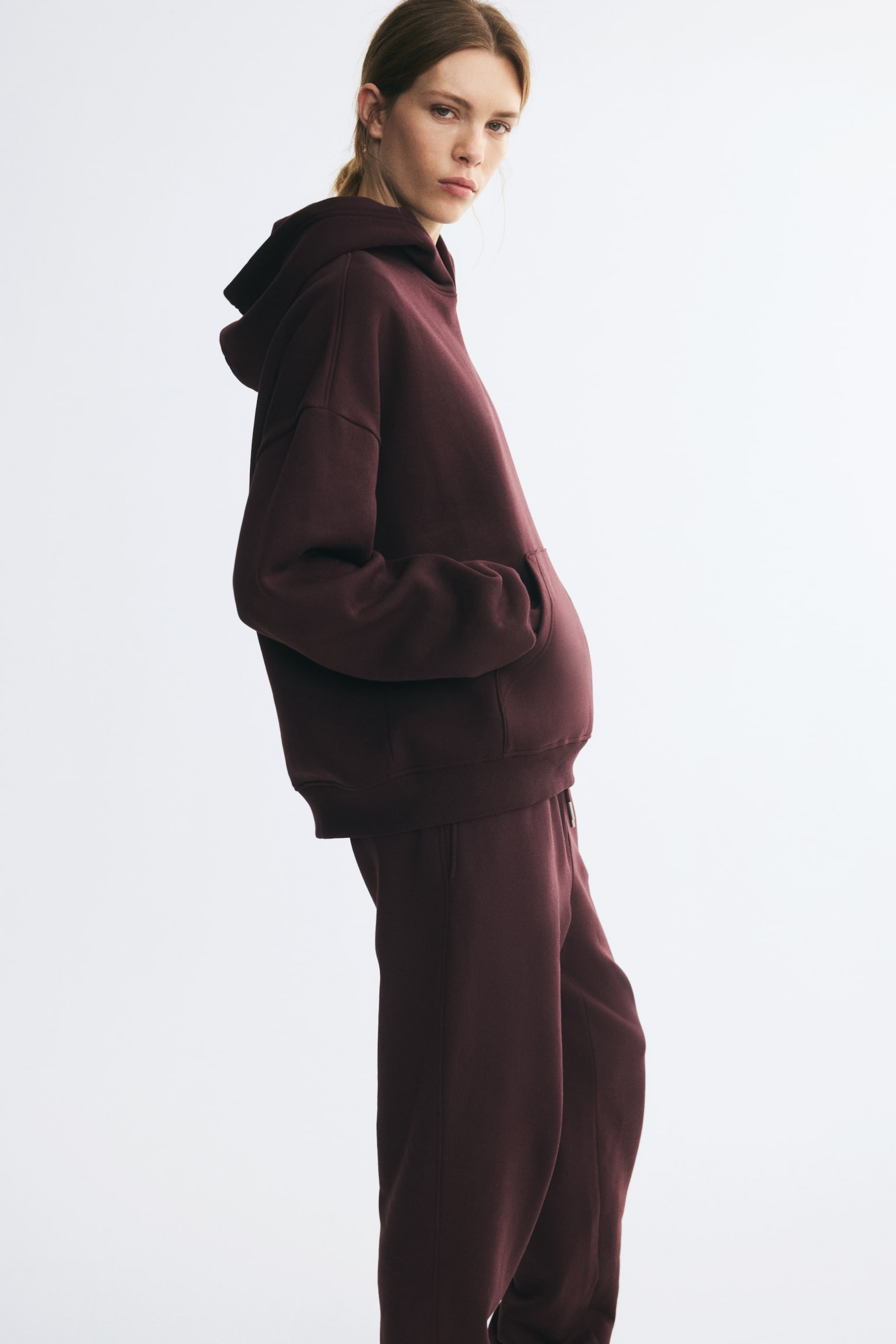 A dark burgundy hoodie in sweat fabric is being worn by the model, who is standing in profile with hands placed inside the kangaroo pocket. The hoodie features low dropped shoulders, a double-layer hood, and ribbed trim, and is paired with matching burgundy sweatpants. - 1