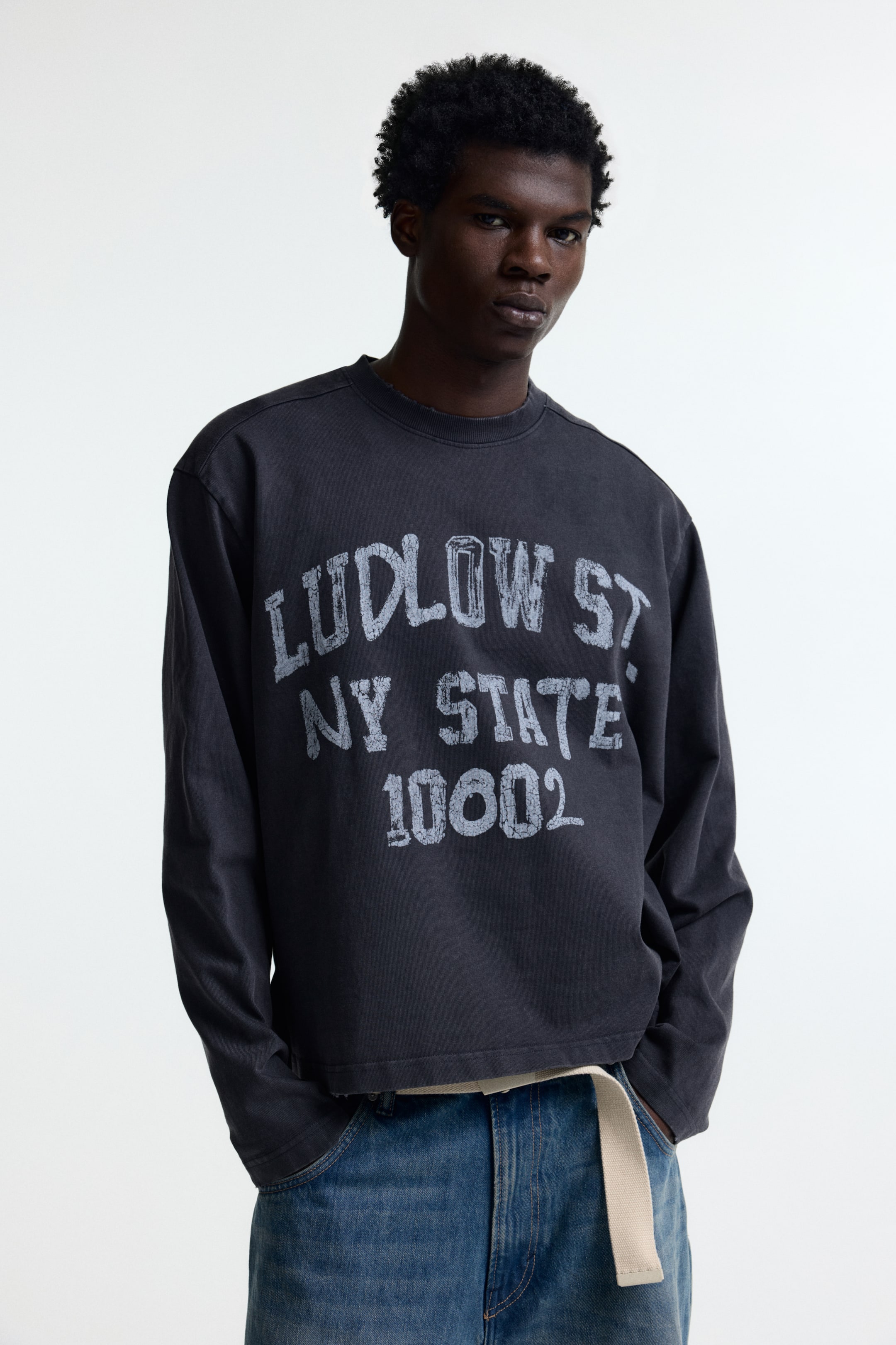 View larger image: A dark gray long-sleeved top is facing forward, featuring a distressed white graphic on the front reading "LUDLOW ST NY STATE 10002". It is designed with a crew neck and a raw-edged, cropped hem, revealing a lighter garment underneath.