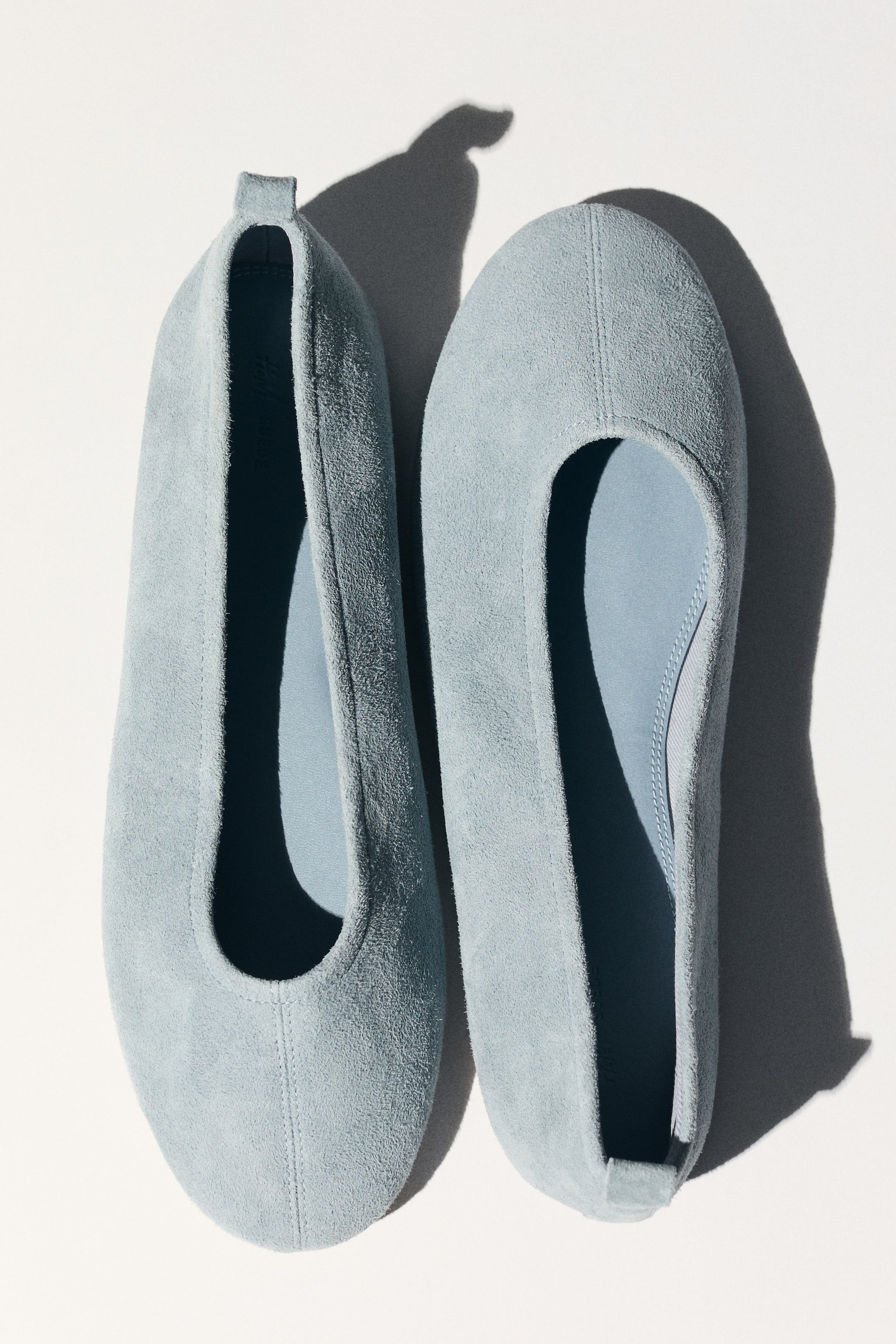 Ampliar la imagen: Dos bailarinas de gamuza azul claro están colocadas una al lado de la otra, presentadas desde un ángulo cenital con las puntas hacia arriba. Cada bailarina cuenta con una costura central prominente que va desde la punta hasta el empeine, su interior está forrado con un material azul más claro a juego, y una pequeña lengüeta es visible en el talón del zapato izquierdo.