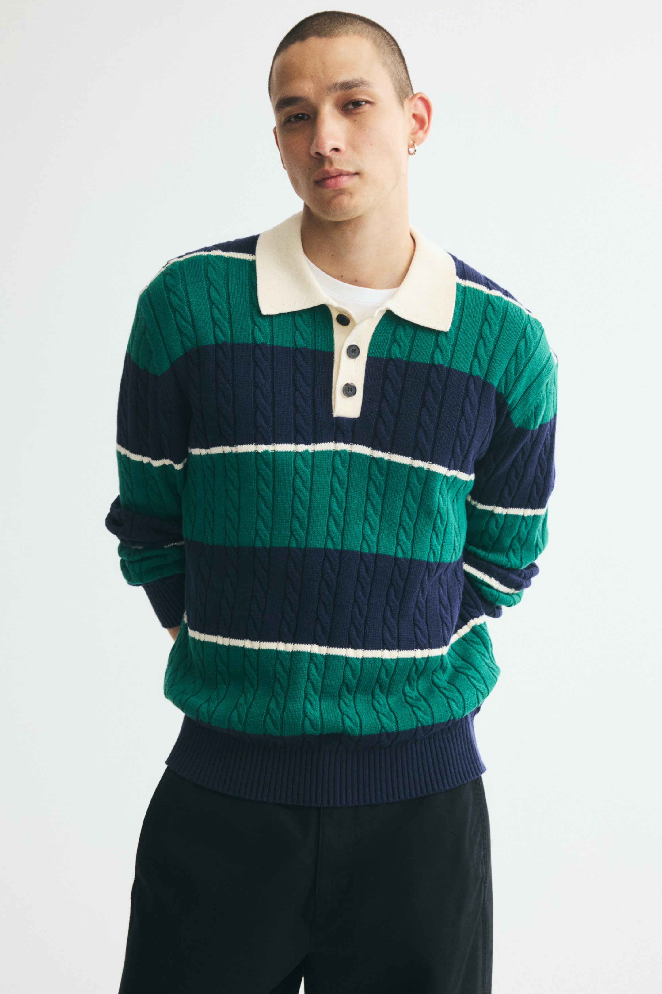 View larger image: A long-sleeve cable-knit polo shirt featuring green, navy, and white horizontal stripes is worn by the model. The shirt has a contrasting cream collar and button placket, and is styled with a layered white T-shirt and black trousers.