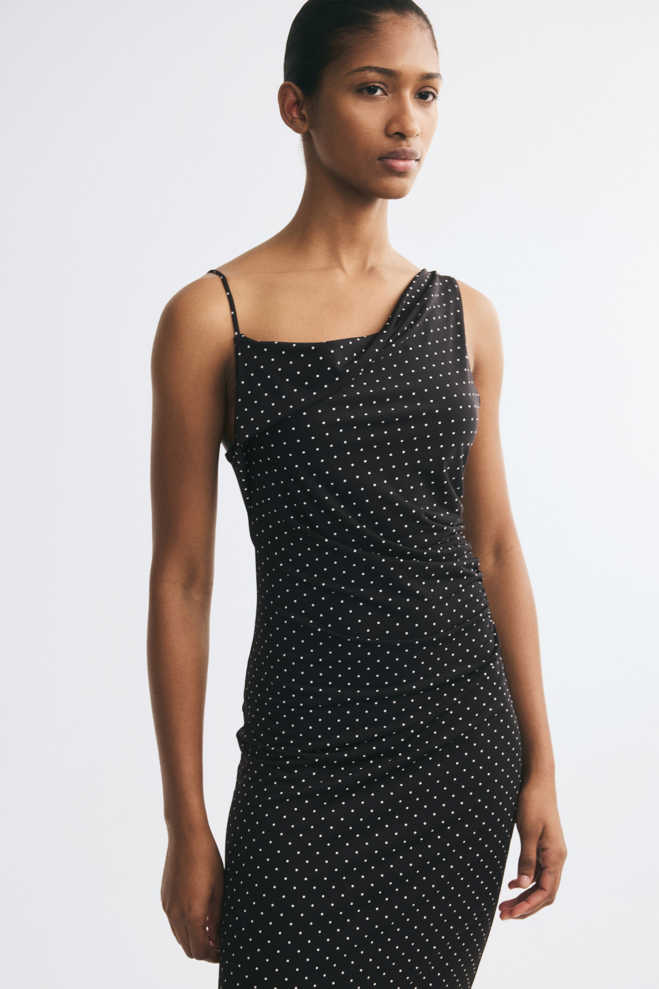Ampliar la imagen: La modelo lleva puesto un vestido negro entallado con lunares blancos, que presenta un escote asimétrico con una tirante delgado en un hombro y una tirante ancho y drapeado en el otro. El vestido tiene fruncido en la cintura que crea un efecto drapeado en la parte frontal.