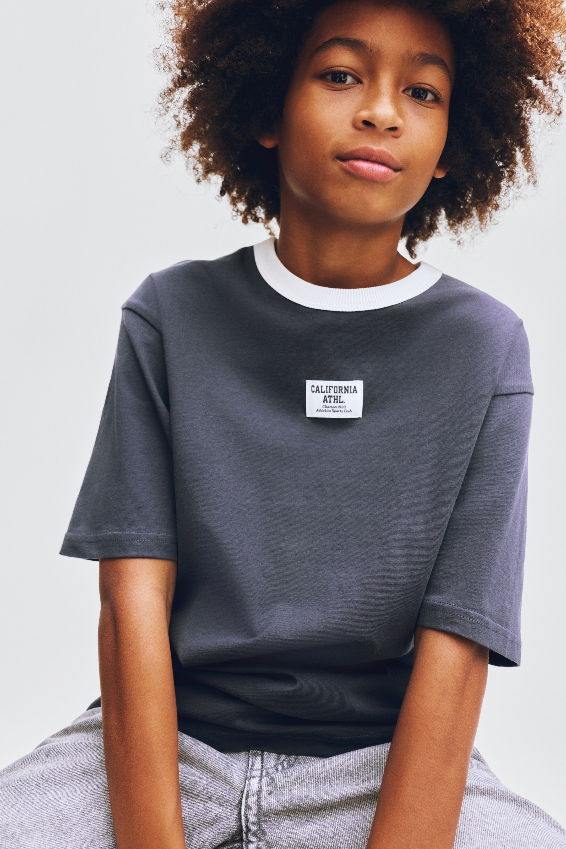 View larger image: A charcoal gray tee showcases a white ribbed crew neck and short sleeves. A rectangular white patch with black text, reading 'CALIFORNIA ATHL. Champs 1992 Athletics Sports Club,' is prominently displayed on the chest.