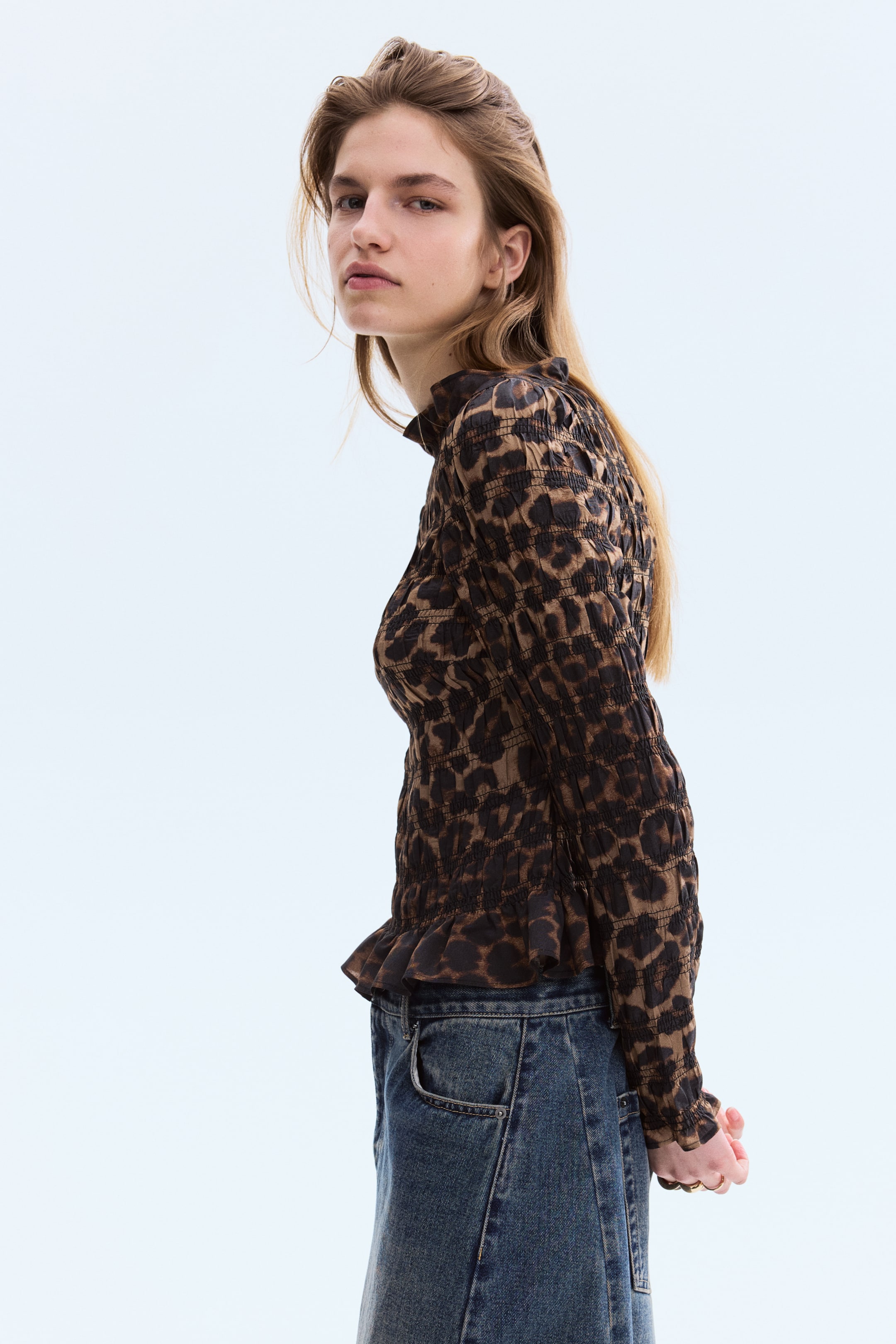 Ampliar la imagen: "Se presenta una blusa con estampado de leopardo en tonos marrón y negro desde un ángulo lateral-frontal. Presenta una textura fruncida en toda la prenda, un cuello alto simulado, mangas largas fruncidas con puños con volantes y un dobladillo peplum con volantes."