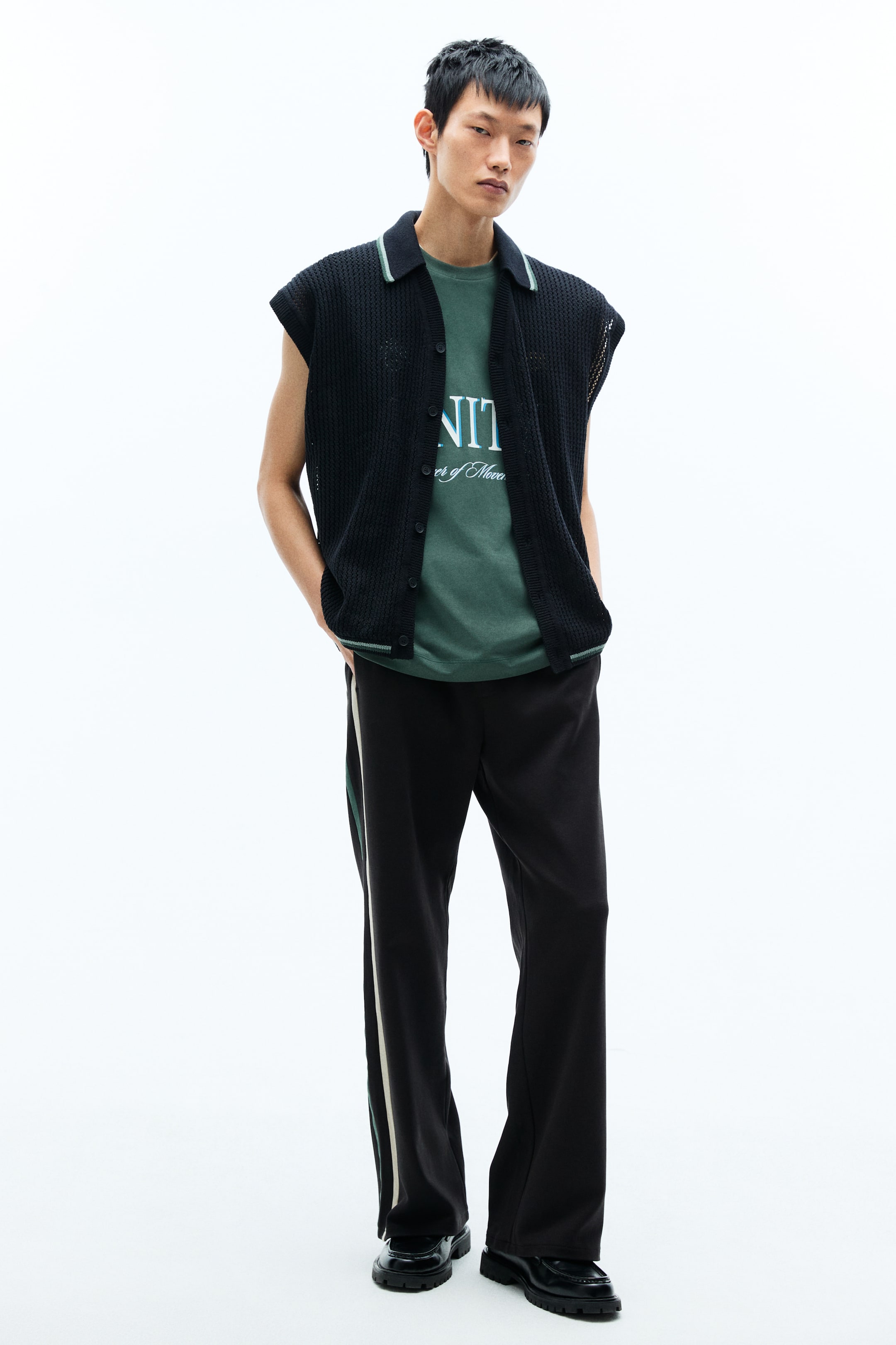 View larger image: A black open-knit sleeveless cardigan is presented front-facing, featuring a polo collar and a ribbed bottom hem detailed with a light green stripe, and a button-up front, worn open over a green T-shirt.
