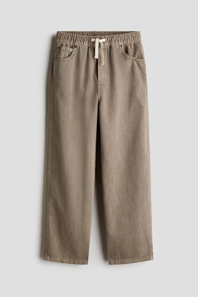 These taupe cotton twill joggers are shown from the front, showcasing a baggy, straight-leg silhouette. They are featuring an elasticated, gathered waistband with an off-white drawstring, a stitched fake fly, and classic five-pocket styling. - 1