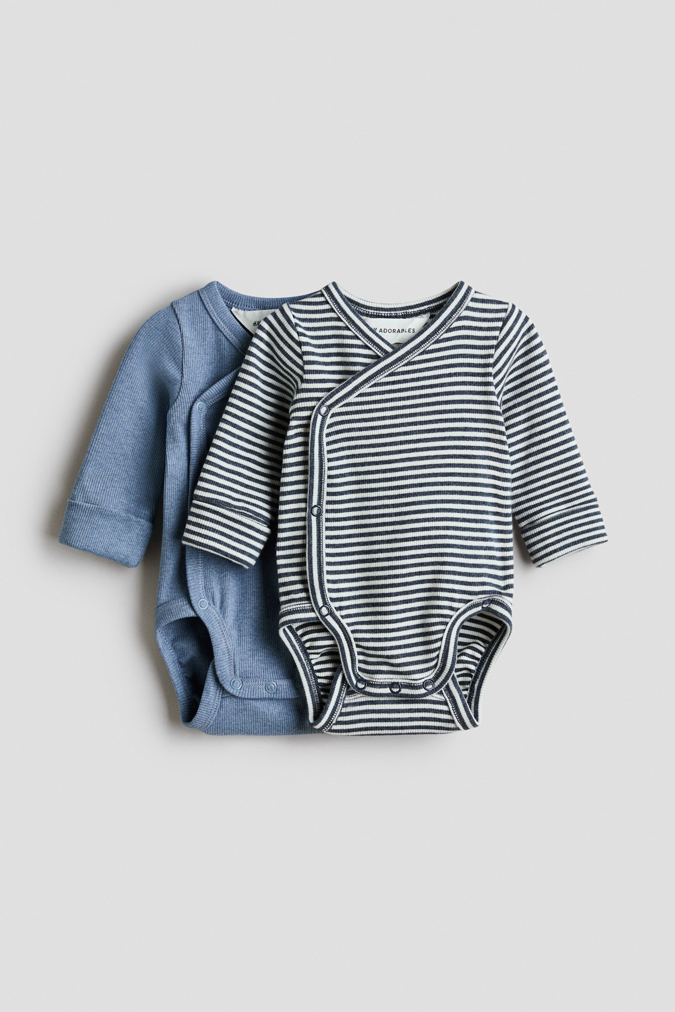 查看大圖: 兩件 H&M Adorables 連身衣平放在一起,白色和深藍色橫條紋羅紋連身衣稍微放在一件純藍色羅紋連身衣的前面。兩件連身衣均採用包裹式設計,側面有按扣,長袖帶摺袖口,底部有雙排按扣。