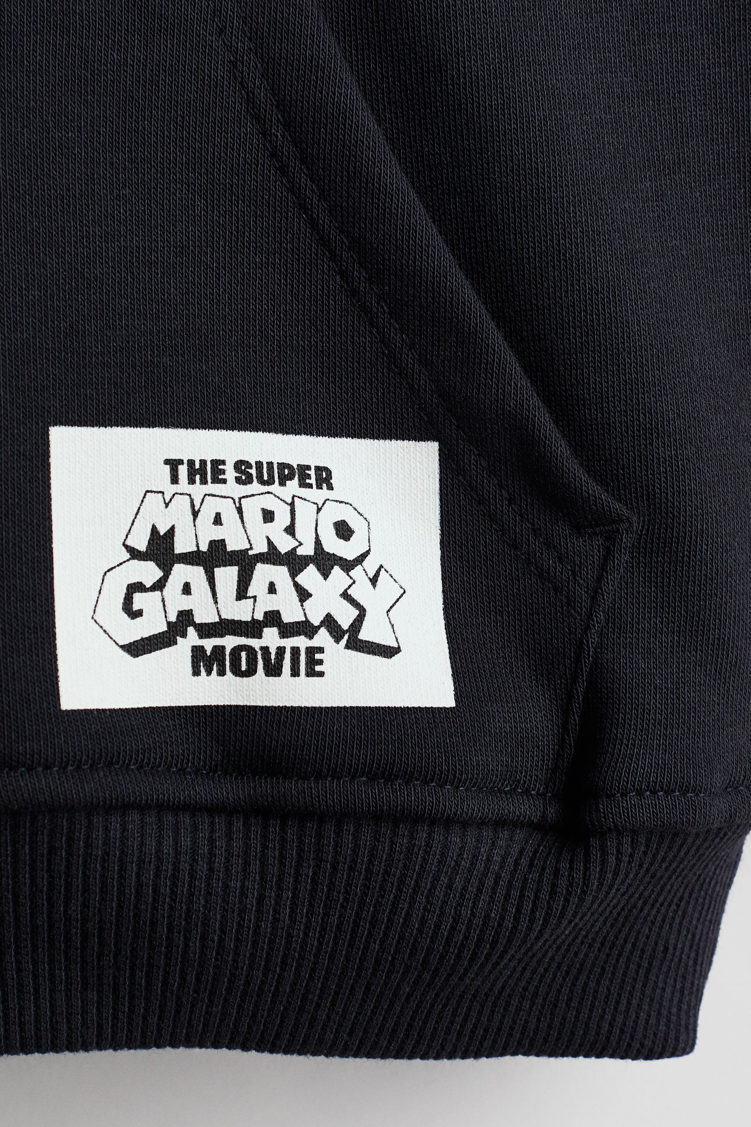 Un primo piano della felpa con cappuccio in pile nero mostra una toppa rettangolare bianca cucita sulla parte anteriore inferiore, sopra lâorlo a costine. Sulla toppa è visibile il testo nero "THE SUPER MARIO GALAXY MOVIE" in un carattere stilizzato, con lâangolo di una tasca a marsupio visibile sulla destra.
