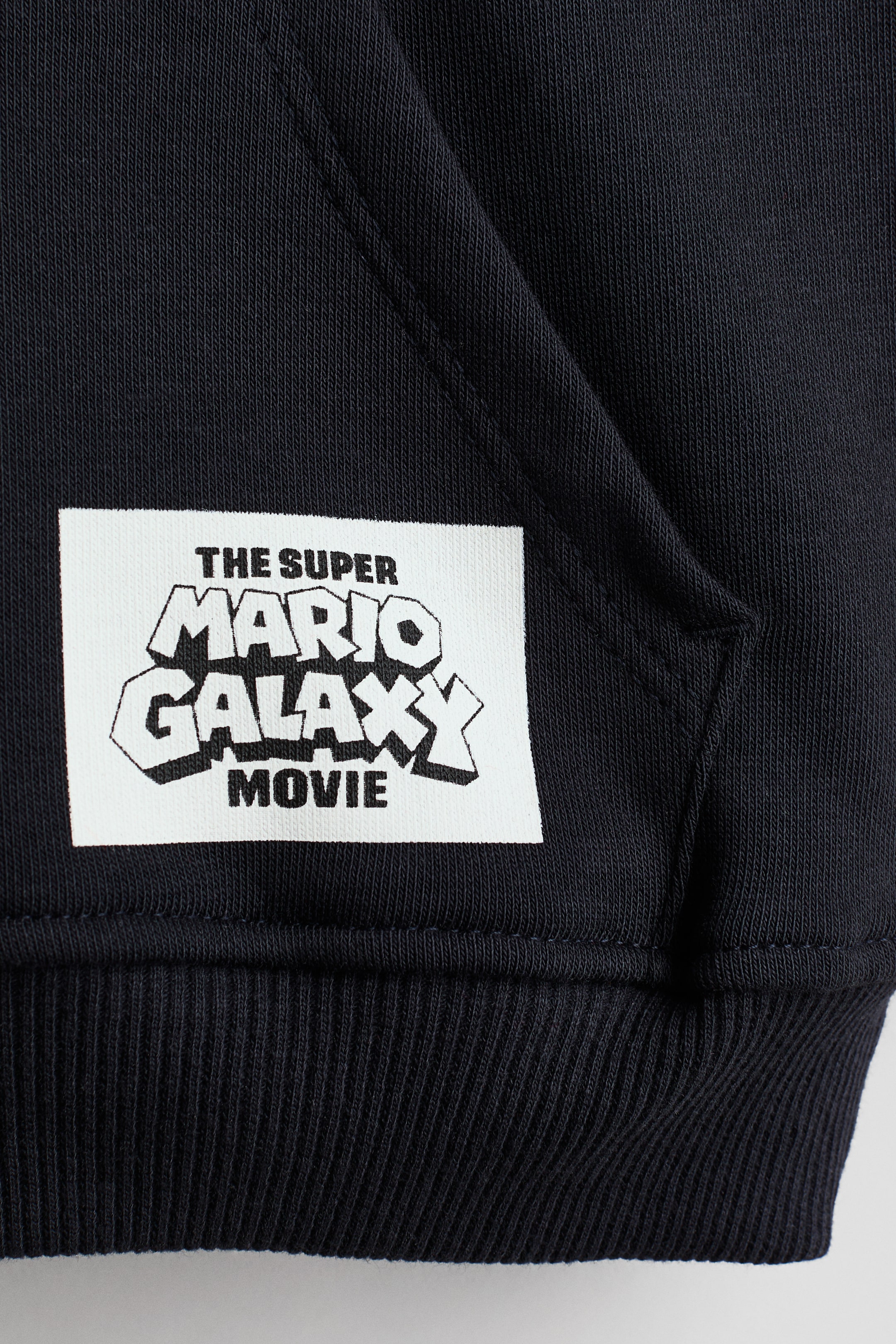 Ver imagem maior: Um close-up do hoodie de polar preto mostra um remendo retangular branco cosido na parte frontal inferior, acima da bainha canelada. O remendo apresenta o texto preto "THE SUPER MARIO GALAXY MOVIE" numa fonte estilizada, com o canto de um bolso canguru visível à direita.