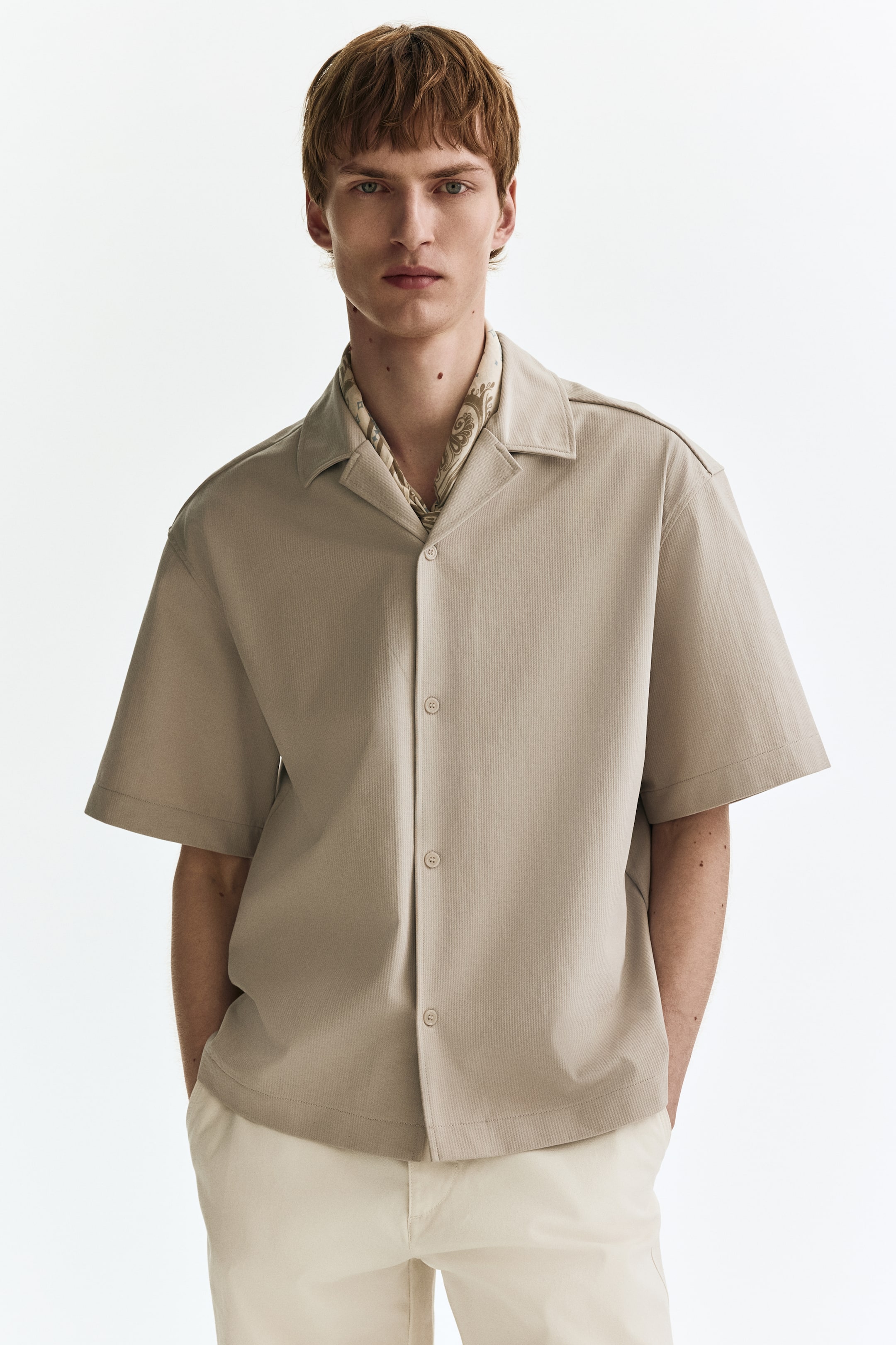 Ampliar la imagen: Una camisa beige de manga corta presenta un ajuste relajado, un cuello abierto tipo spread, cierre frontal con botones y un fino ribeteado vertical, con un pañuelo estampado usado debajo del cuello.
