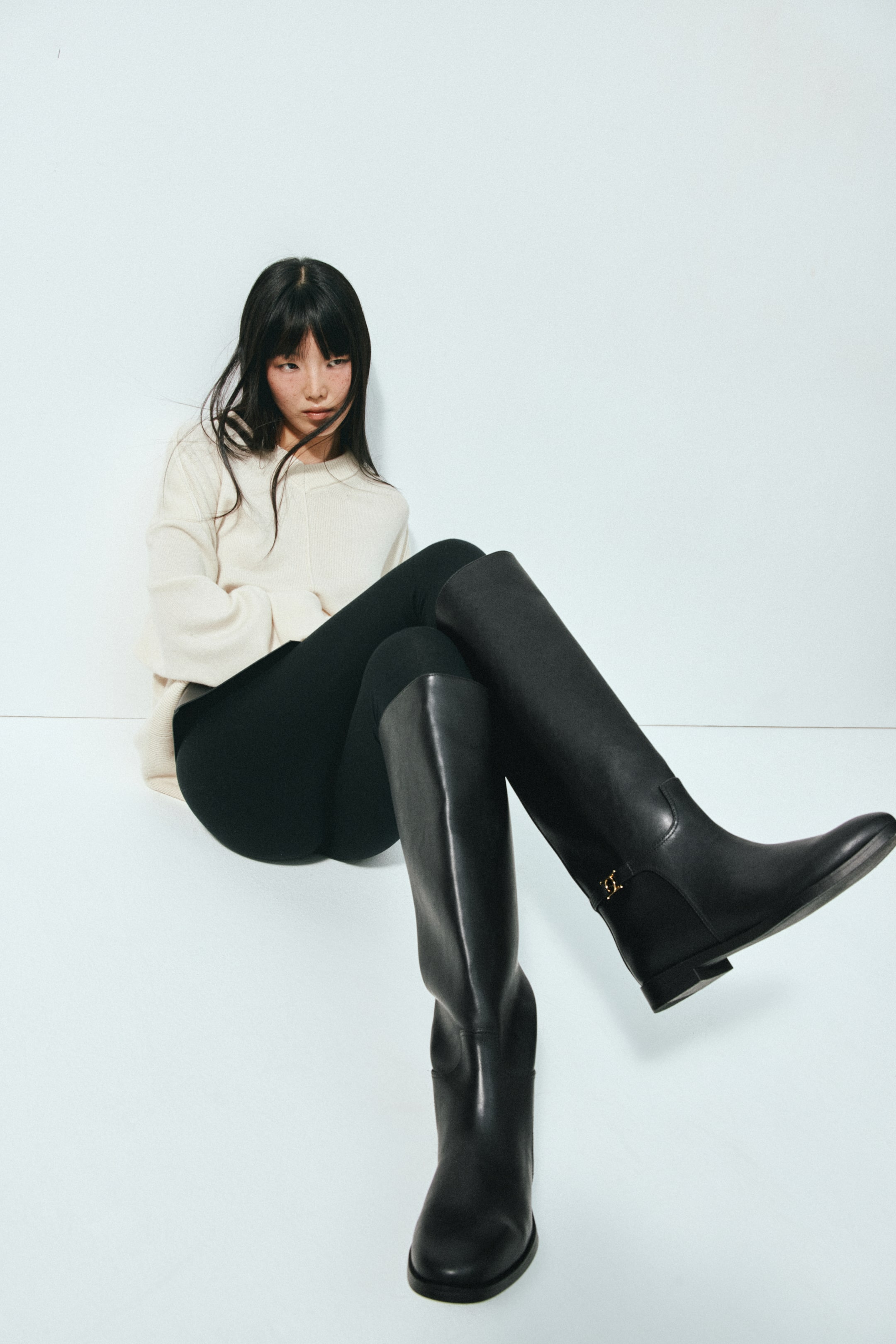 Agrandir l'image: Des bottes noires montantes jusqu’au genou, dotées d’orteils arrondis, de petits talons blocs et d’une bride décorative avec boucle métallique autour de la cheville, sont portées par le mannequin. Les bottes sont associées à un legging noir et à un pull crème, avec une jambe relevée pour mettre en valeur le détail de la cheville.