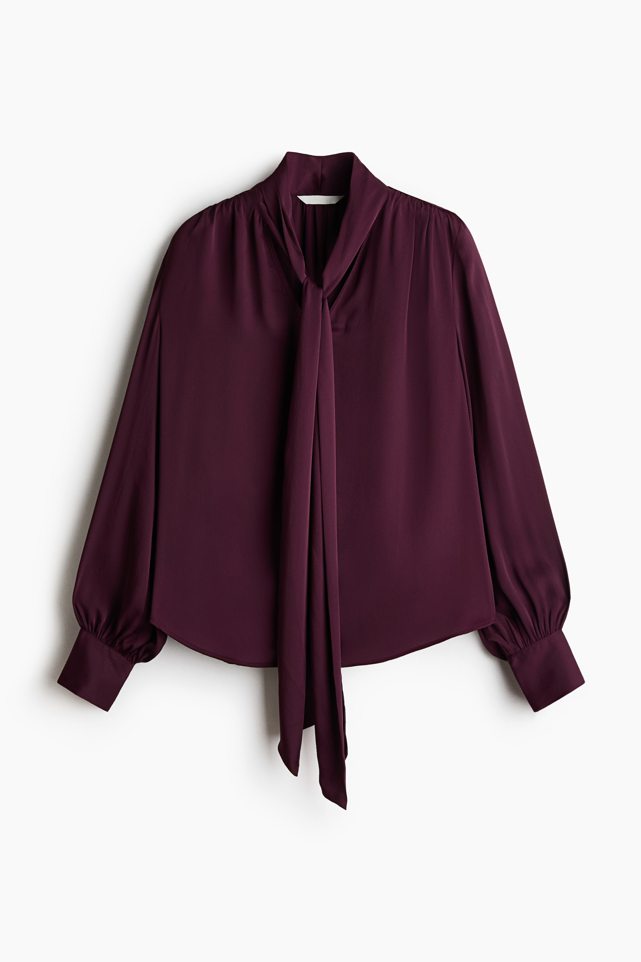 View larger image: A maroon blouse features a high neck with a long tie detail knotted at the front, voluminous long sleeves gathered at the buttoned cuffs, and a curved hemline.