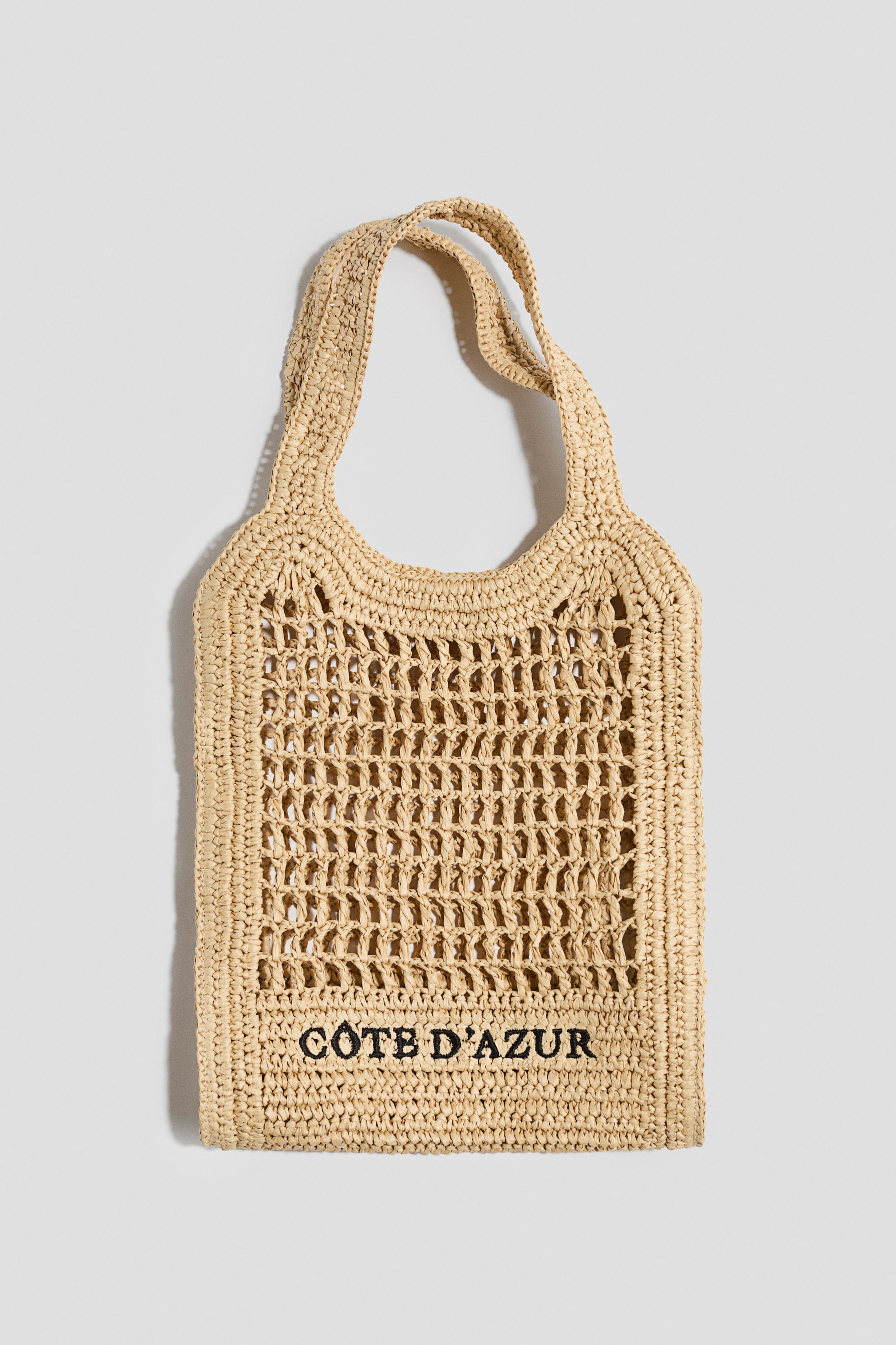 View larger image: A woven raffia tote bag is showcased from the front, highlighting its open-netted body with tightly woven borders and handles. The words "CÔTE D'AZUR" are embroidered in black on the lower front.