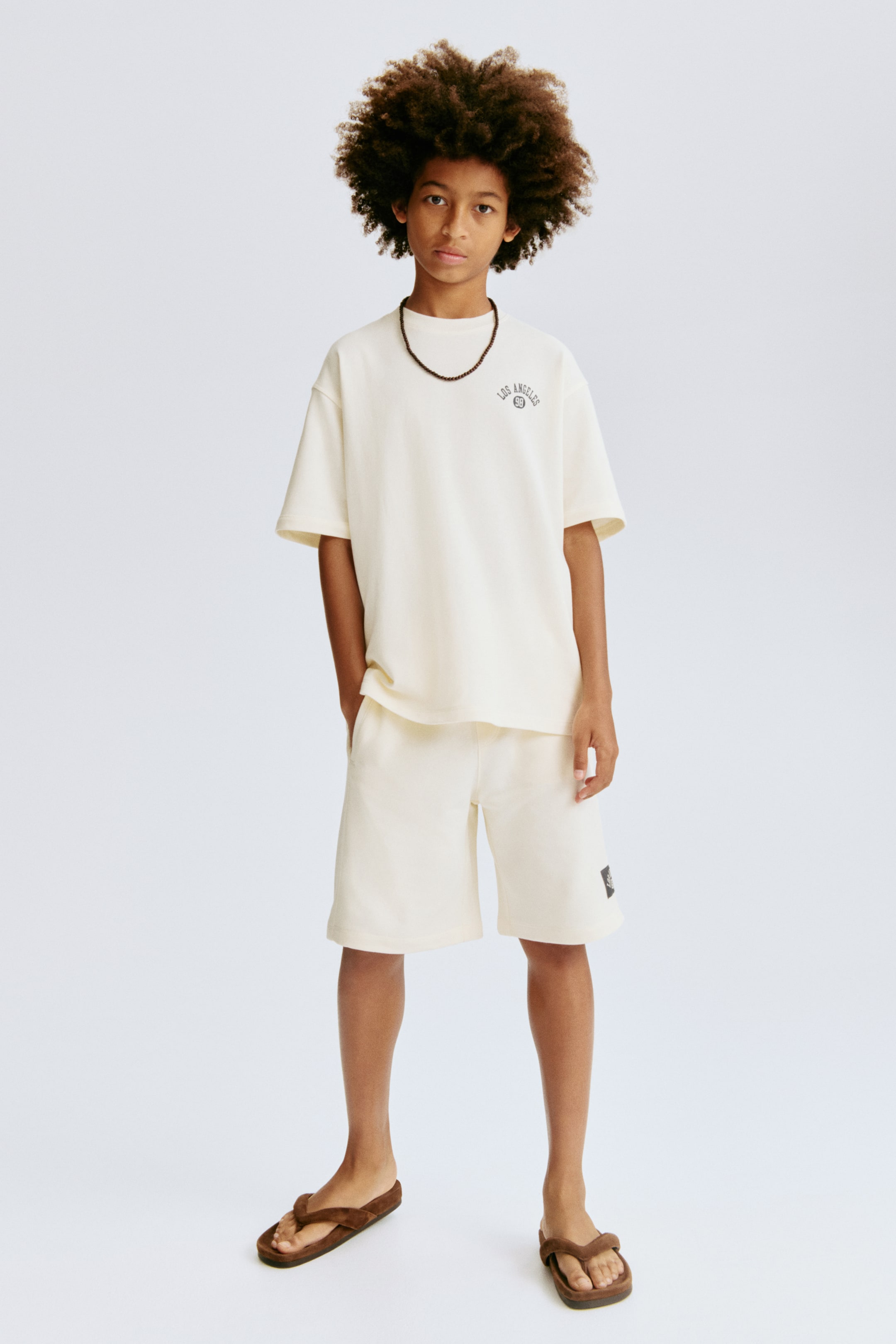 Ver imagen más grande: Un conjunto de camiseta de manga corta y pantalones cortos a juego en color crema se presenta desde un ángulo frontal. La camiseta de corte relajado muestra un pequeño gráfico circular oscuro en el pecho izquierdo, y los pantalones cortos exhiben un pequeño parche cuadrado oscuro en la pierna izquierda.