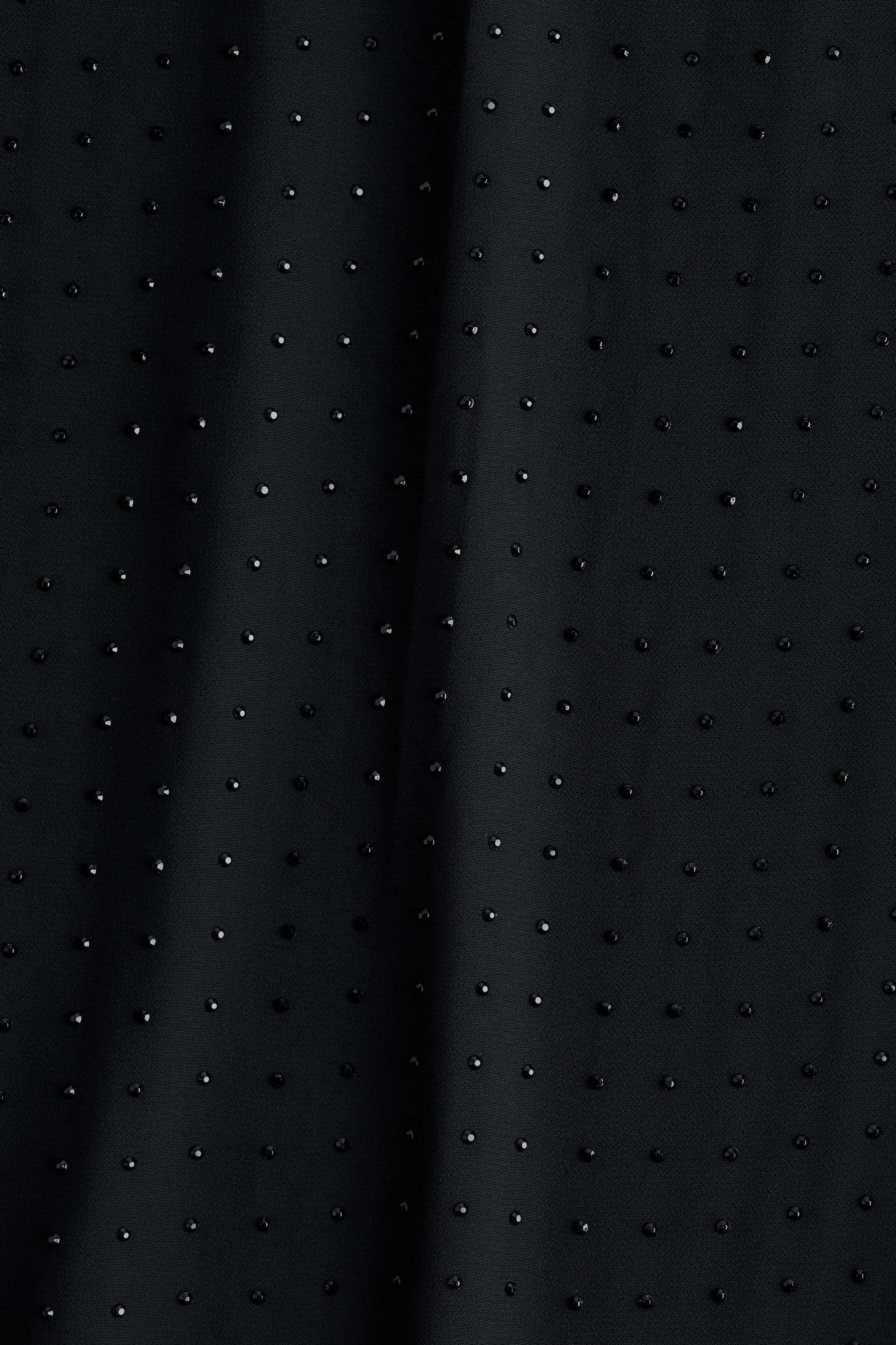Ampliar la imagen: Una tela negra está cubierta con cuentas negras pequeñas y facetadas, espaciadas uniformemente, creando una textura punteada. Un pliegue vertical suave aparece en el lado izquierdo de la tela.