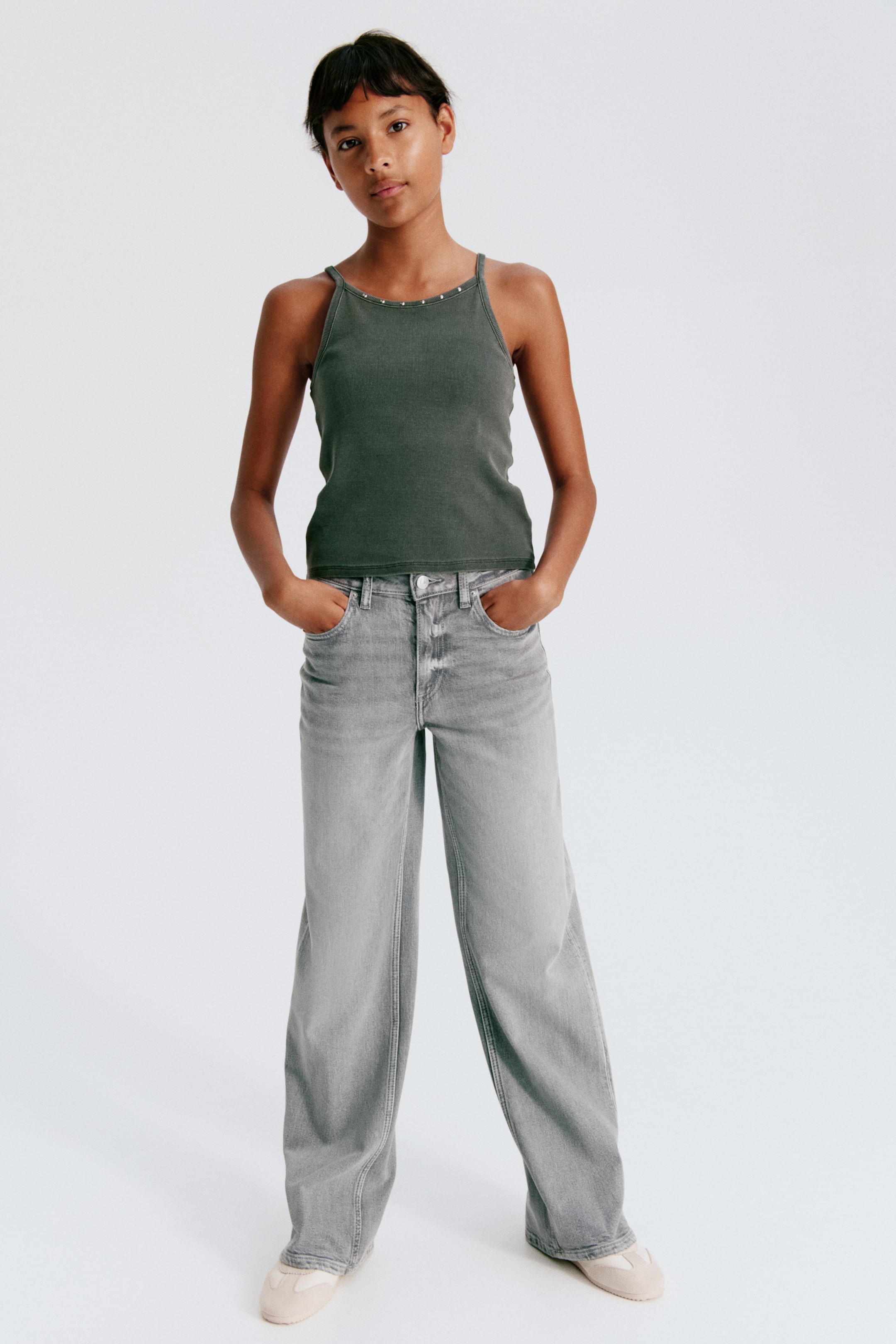 Größeres Bild ansehen: Ein dunkelgrünes Neckholder-Top und hellgraue Jeans mit weitem Bein sind aus frontaler Perspektive zu sehen. Das Top verfügt über schmale Spaghettiträger mit kleinen metallischen Nieten entlang des Ausschnitts, während die lässig geschnittene Jeans eine verwaschene Waschung und den klassischen Five-Pocket-Stil aufweist.