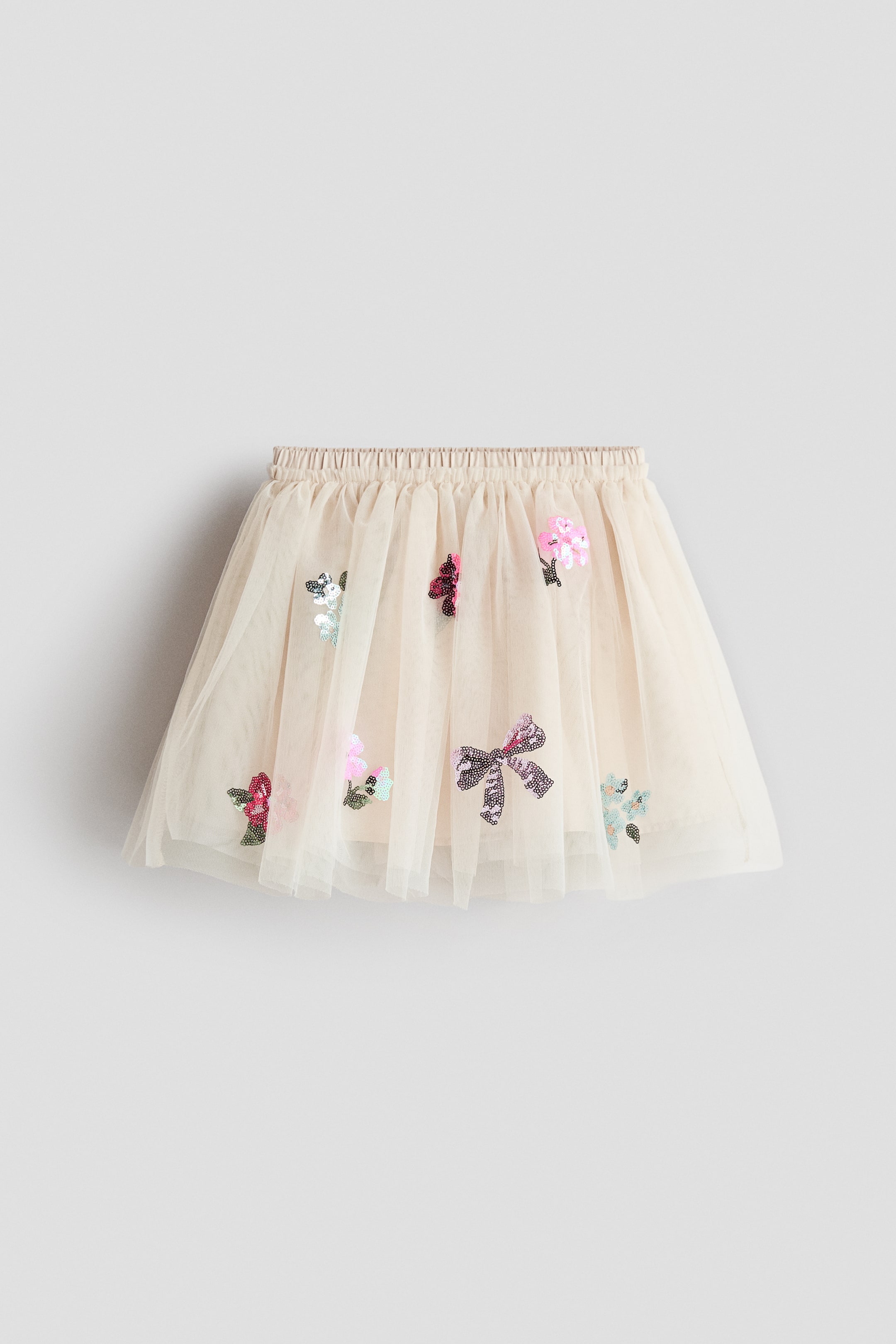 View larger image: A cream-coloured, flared skirt is showcasing sheer tulle with an elasticated, gathered waist. It is featuring scattered sequin motifs of flowers and bows in various colours, and an opaque lining is visible underneath.
