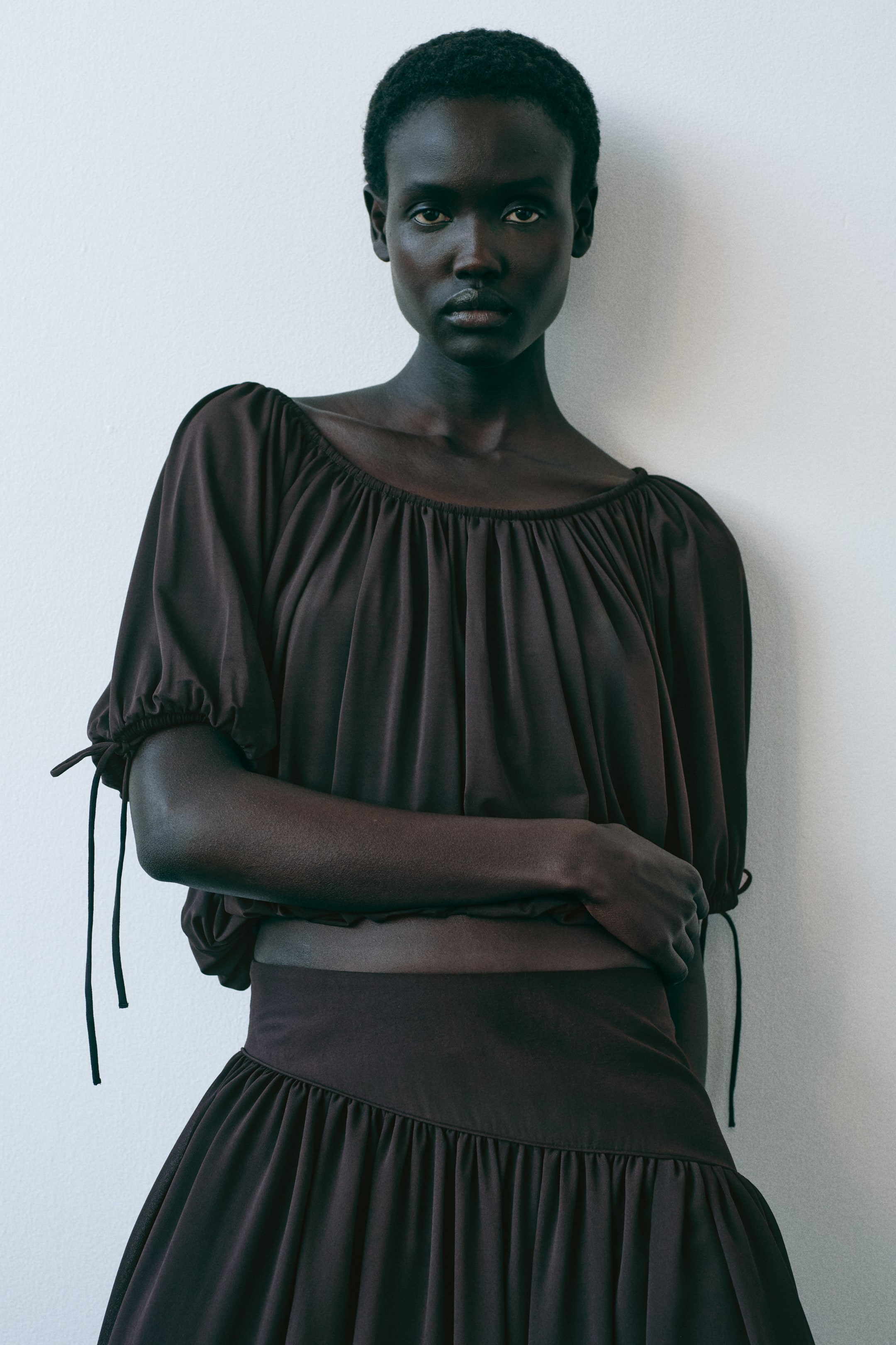 View larger image: Presenting a front-facing dark brown two-piece set, this ensemble features a top with a wide, gathered neckline and puffed short sleeves accented with tie details. Complementing the top is a matching skirt, designed with a wide waistband and a tiered, gathered silhouette. The set is styled together, showcasing a bare midriff.