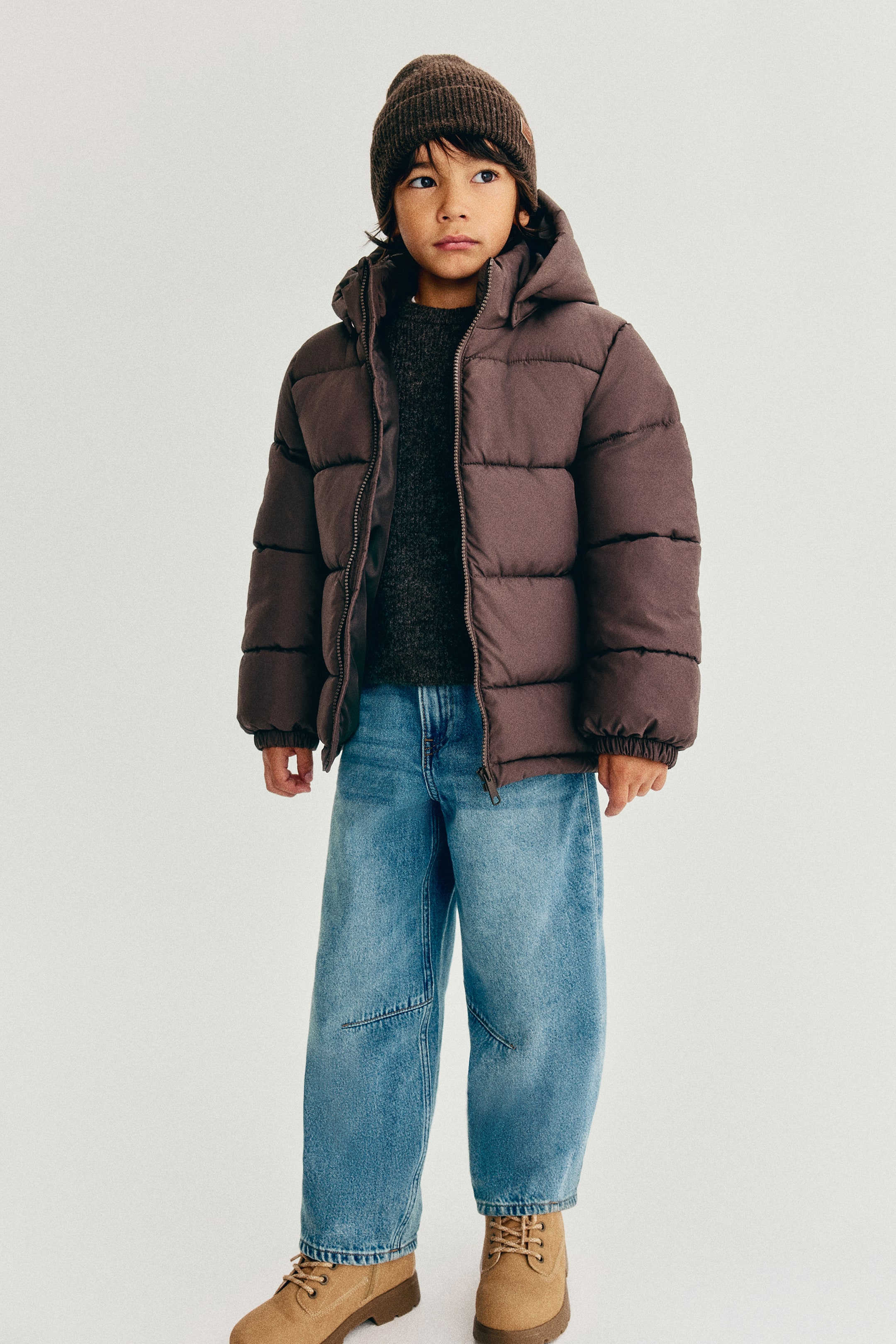 Größeres Bild anzeigen: Eine braune Steppjacke wird von vorne gezeigt und verfügt über horizontale Steppbahnen, eine Kapuze, einen Frontreißverschluss und elastische Bündchen. Die Jacke wird über einem dunkelgrauen Strickpullover getragen und mit hellblauen Jeans in heller Waschung kombiniert.