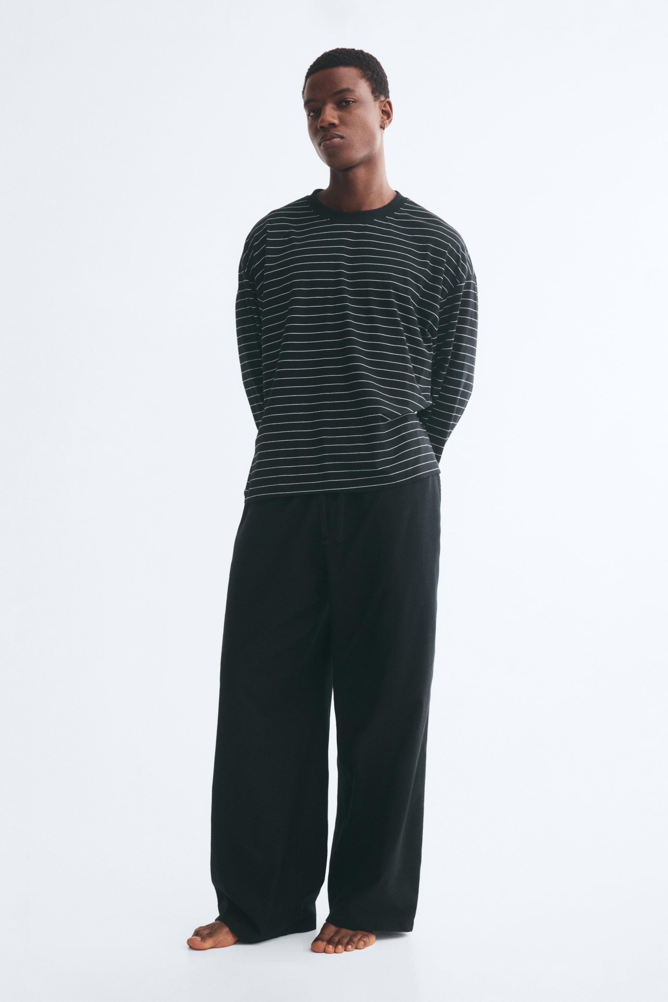 Grotere afbeelding bekijken: Een loungewear-set bestaande uit een zwart-wit horizontaal gestreepte jersey top met lange mouwen en een losse zwarte broek wordt gedragen door het model. De top heeft een ronde hals met ribboord en verlaagde schoudernaden, terwijl de broek is voorzien van een elastische tailleband met trekkoord.