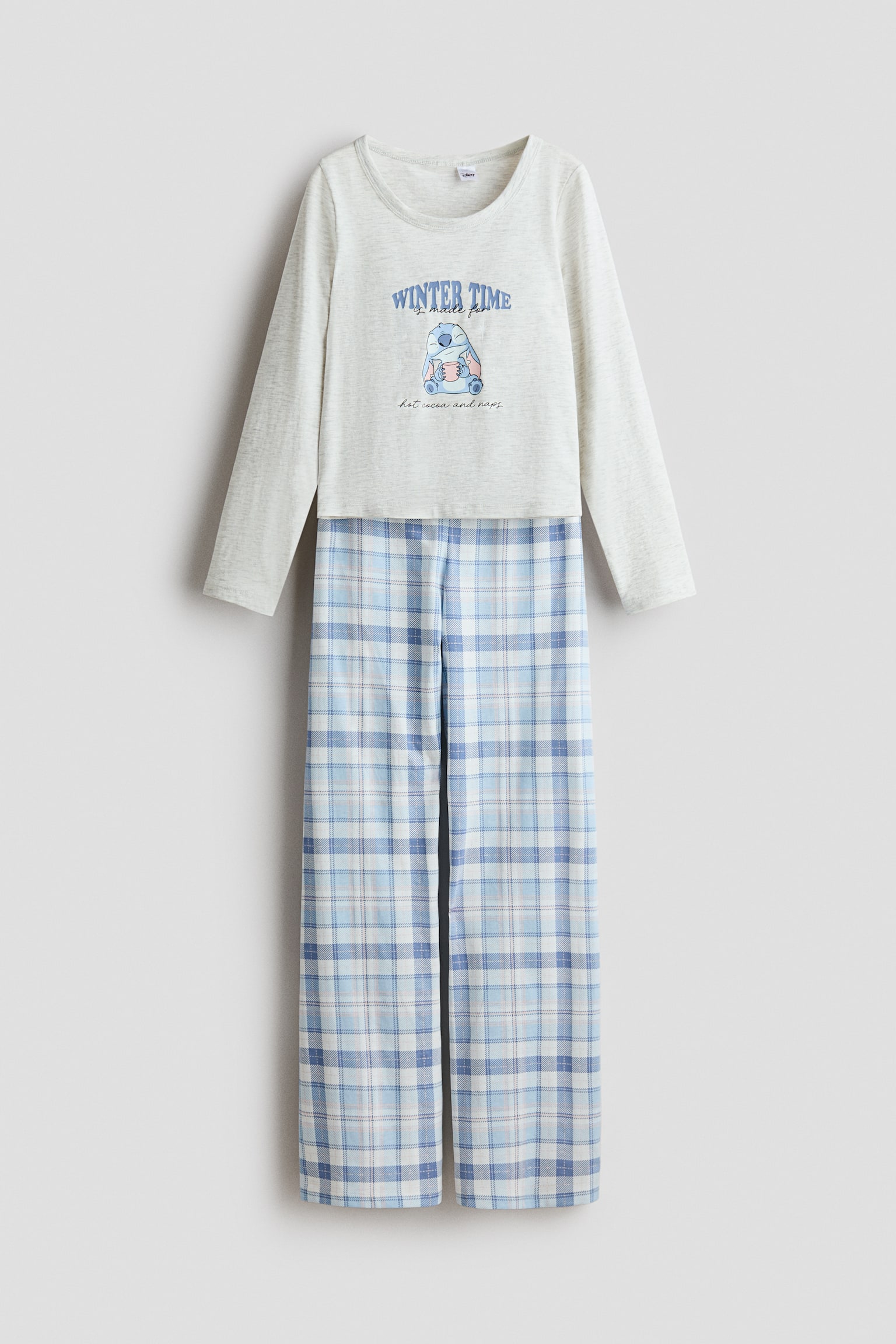 A two-piece pajama set features a light gray heathered long-sleeve top and coordinating blue and white plaid pants, shown from the front. The crew-neck top displays a central graphic of a cartoon character holding a mug with the text "WINTER TIME is made for hot cocoa and naps," while the straight-leg pants showcase an all-over plaid pattern.
