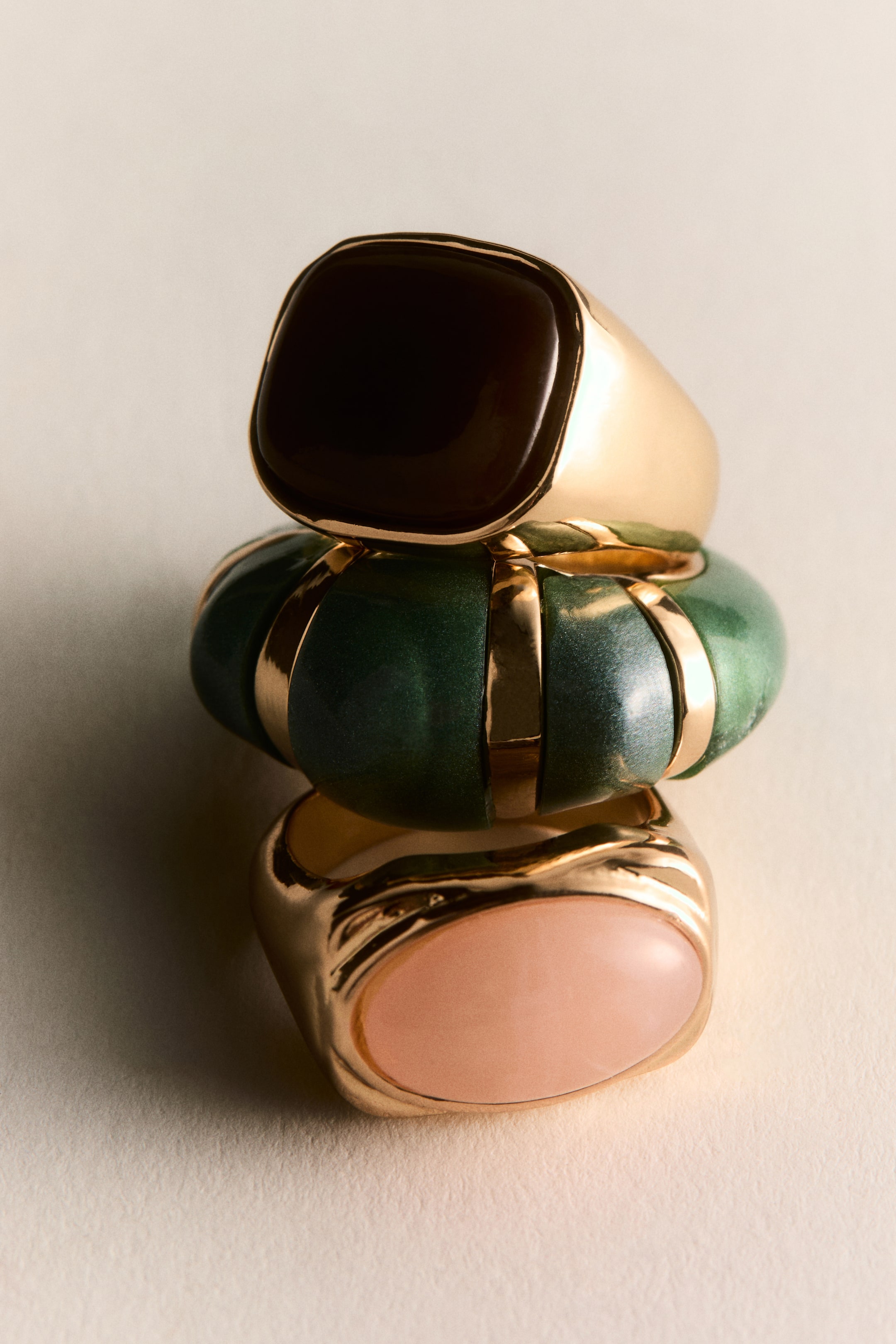 Agrandir l'image: Trois bagues sont empilées verticalement, légèrement inclinées vers la droite. La bague supérieure présente une pierre carrée brun foncé sertie dans un anneau doré. La bague du milieu est large, bombée et verte, ornée horizontalement de plusieurs bandes dorées, tandis que la bague inférieure arbore une pierre ovale rose clair sertie dans un anneau doré.