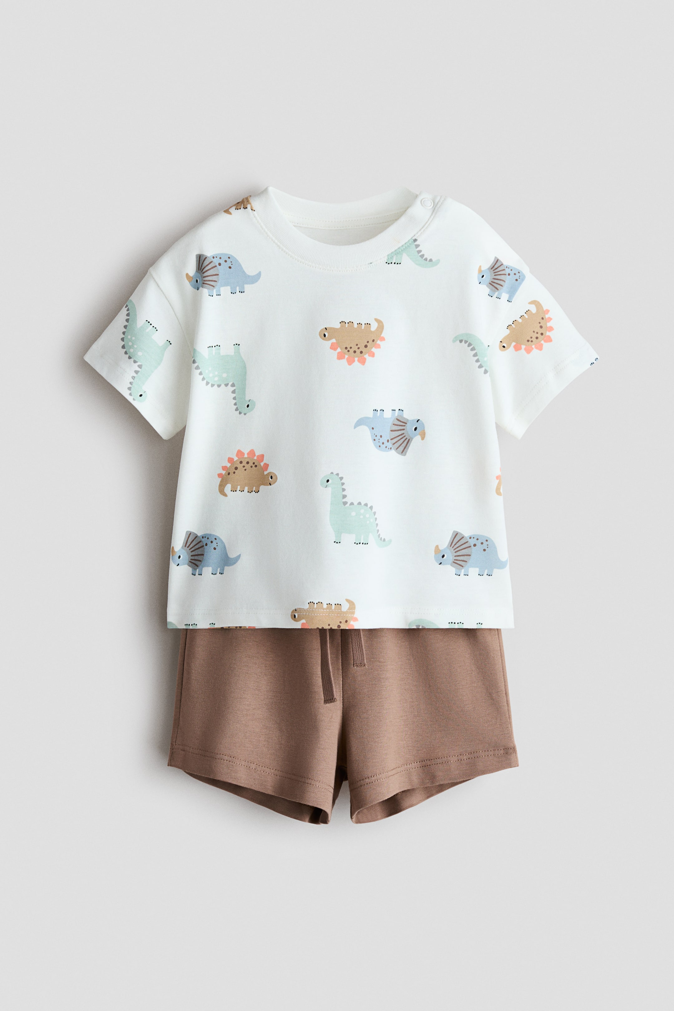 View larger image: A 2-piece cotton set na may kasamang puting t-shirt, ipinapakita mula sa harap, na may all-over print ng makukulay na dinosaurs, bilog na leeg, maikling manggas, at snap buttons sa isang balikat. Ang brown na shorts, dinisenyo na may elasticated na baywang at drawstring, ay nakalayer sa ilalim ng t-shirt.