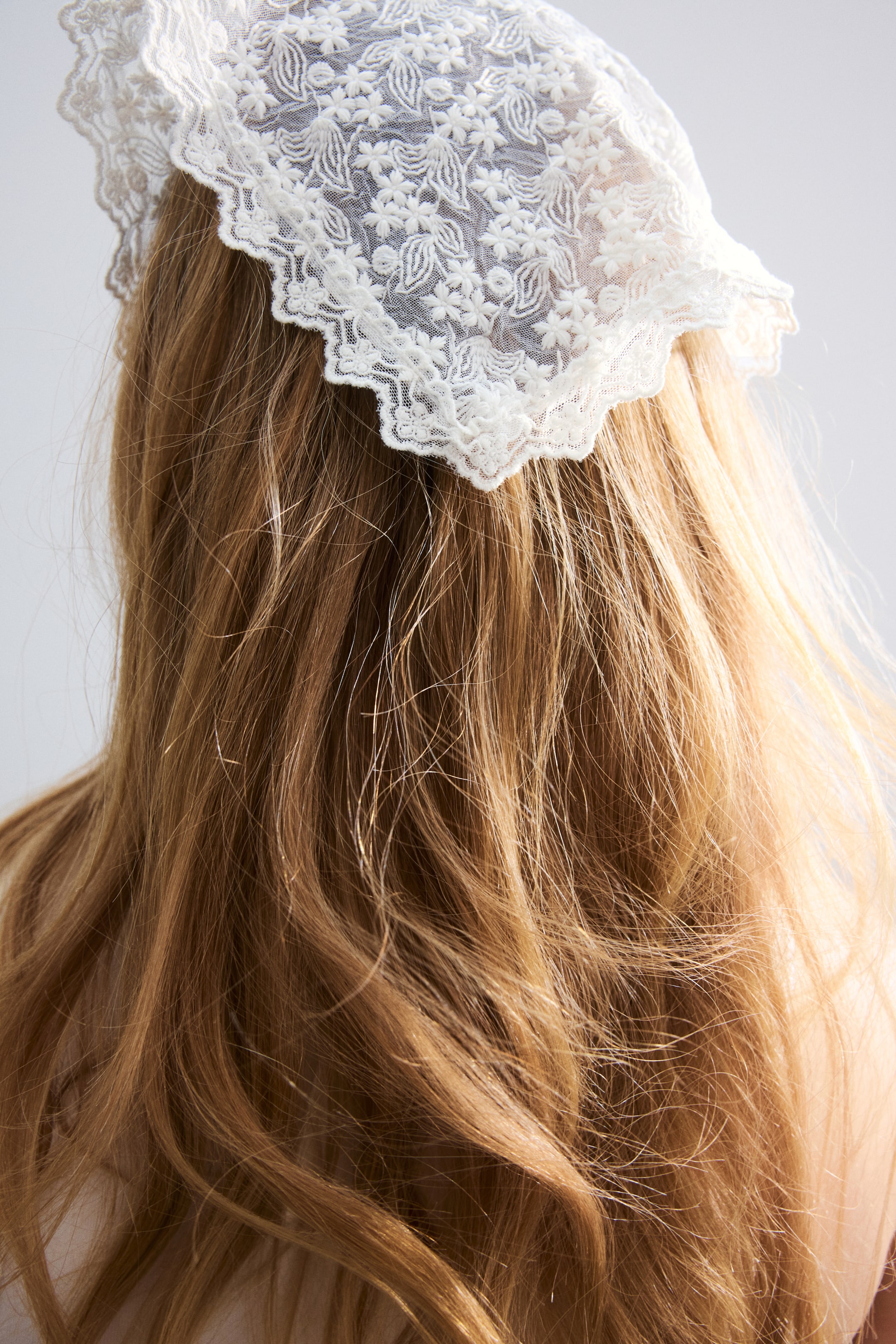 View larger image: A white lace hair accessory adorns light brown hair, viewed from the back, showcasing detailed floral embroidery and a scalloped, ruffled edge.