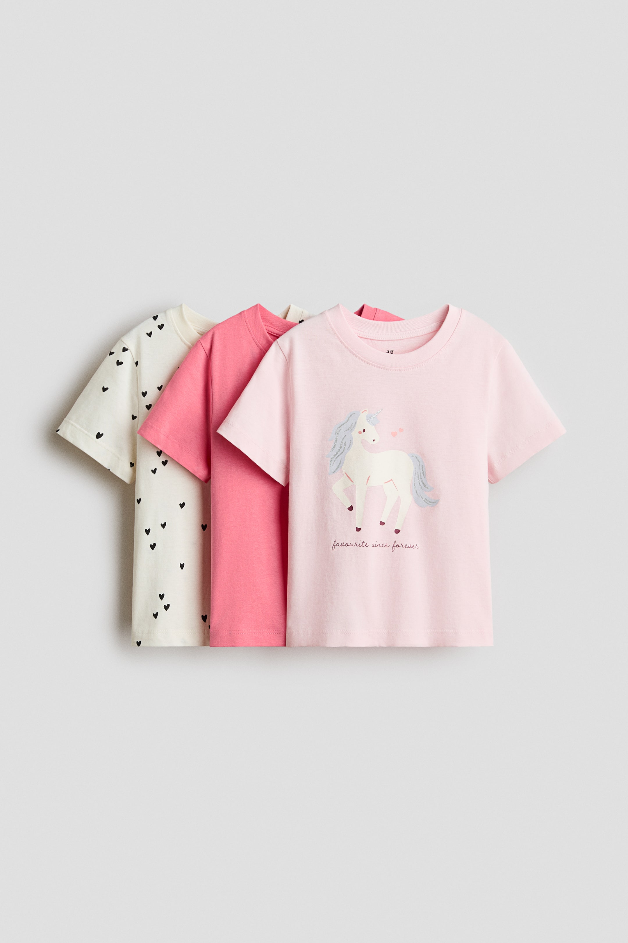Voir une image plus grande: Trois T-shirts en jersey de coton doux imprimé affichent leurs devants. Un T-shirt rose pâle, avec un motif de licorne et du texte, est positionné à l'avant, tandis qu'un T-shirt corail uni et un T-shirt blanc cassé avec un imprimé intégral de cœurs noirs sont superposés derrière. Tous les T-shirts présentent des encolures côtelées et des épaules légèrement tombantes.
