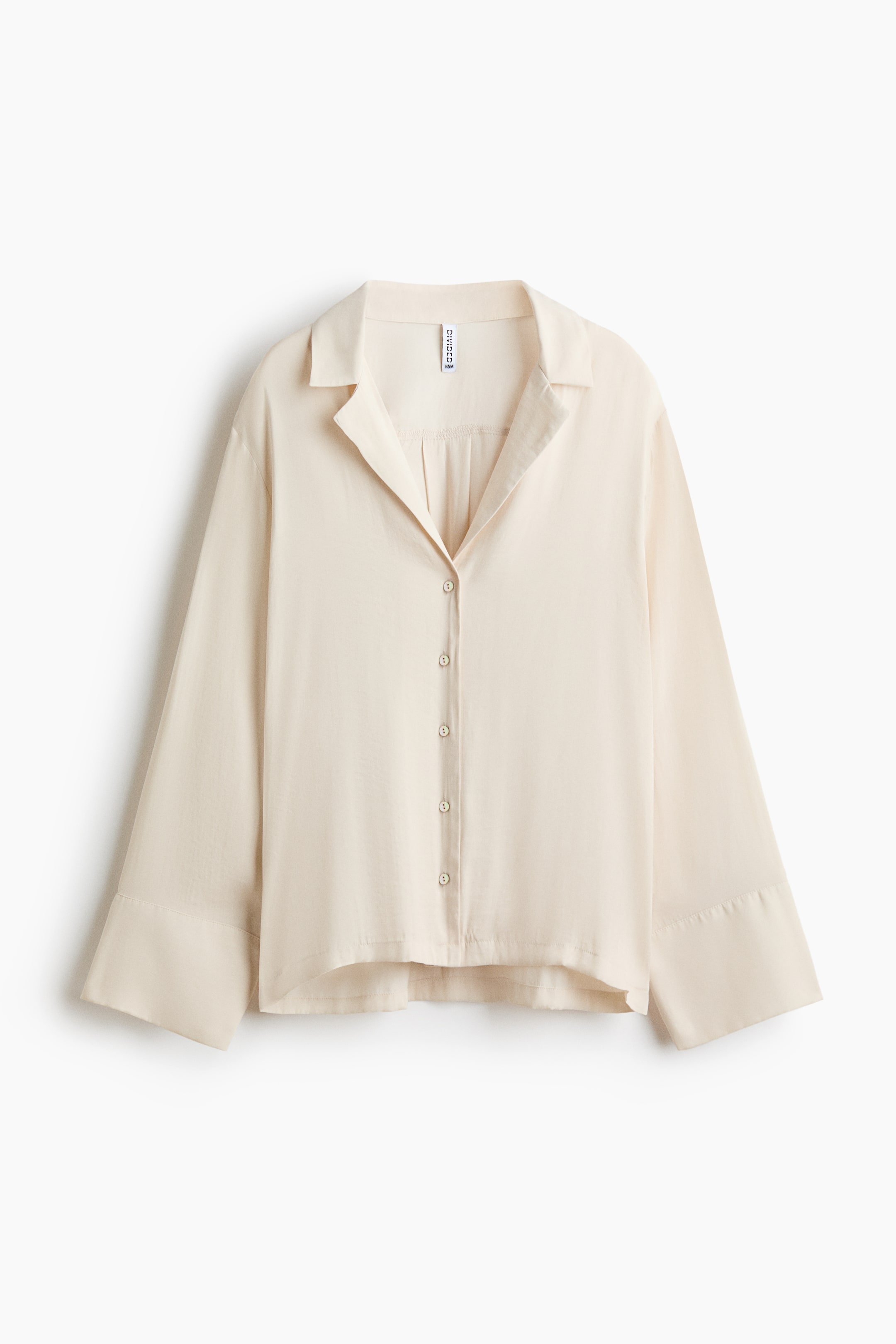 Ver imagen más grande: Una blusa de color crema muestra su parte frontal, con un cuello en punta abierto, una tapeta completa de botones con seis pequeños botones redondos de color crema y sutiles pliegues verticales debajo del cuello. Mangas largas y anchas con puños amplios y un dobladillo curvado completan su diseño relajado.