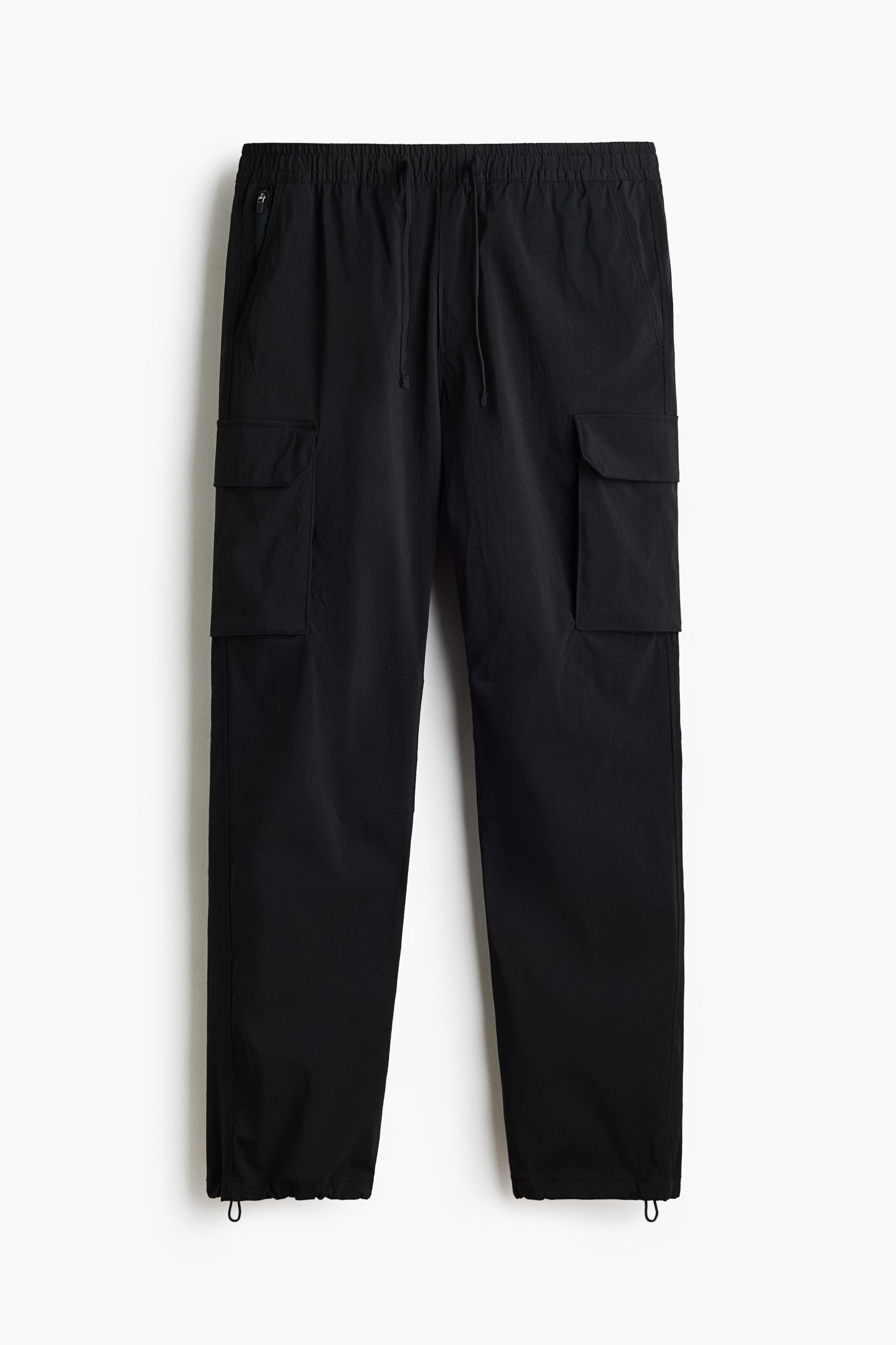 View larger image: Black cargo trousers are displayed from the front. They feature an elasticated waistband with a visible drawstring. Two angled side pockets are shown, one with a zip, and two large flap cargo pockets appear on the thighs, while the cuffs include adjustable drawstrings.