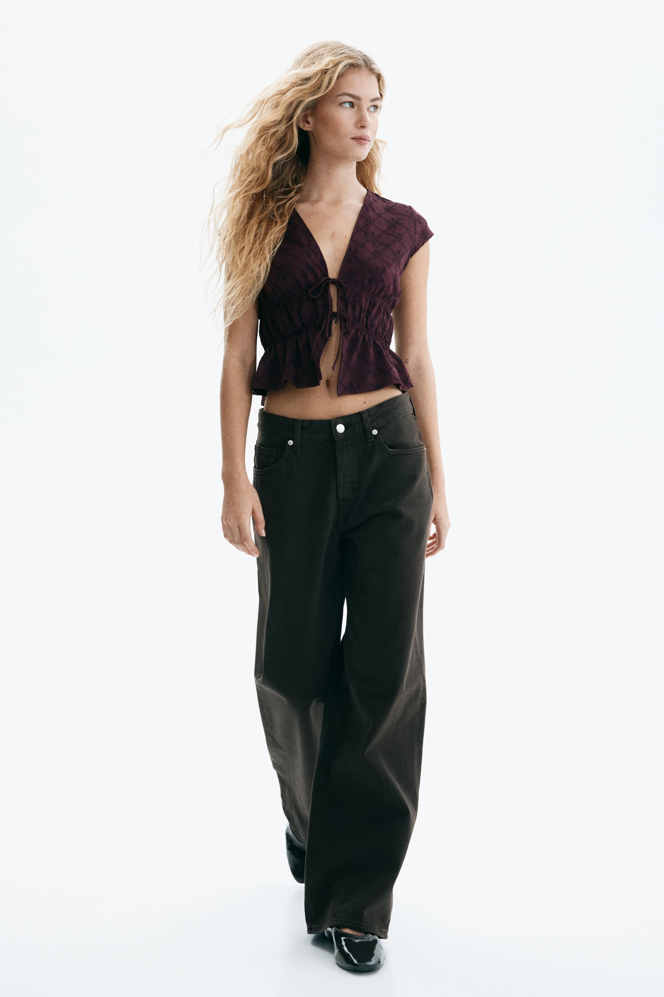 Xem ảnh lớn hơn: Áo croptop tay ngắn màu tím đậm có họa tiết hình học tinh tế, cổ chữ V và chi tiết buộc phía trước tạo gấu xếp nếp. Áo được kết hợp với quần ống rộng màu xám đậm, toàn bộ trang phục hướng về phía trước.