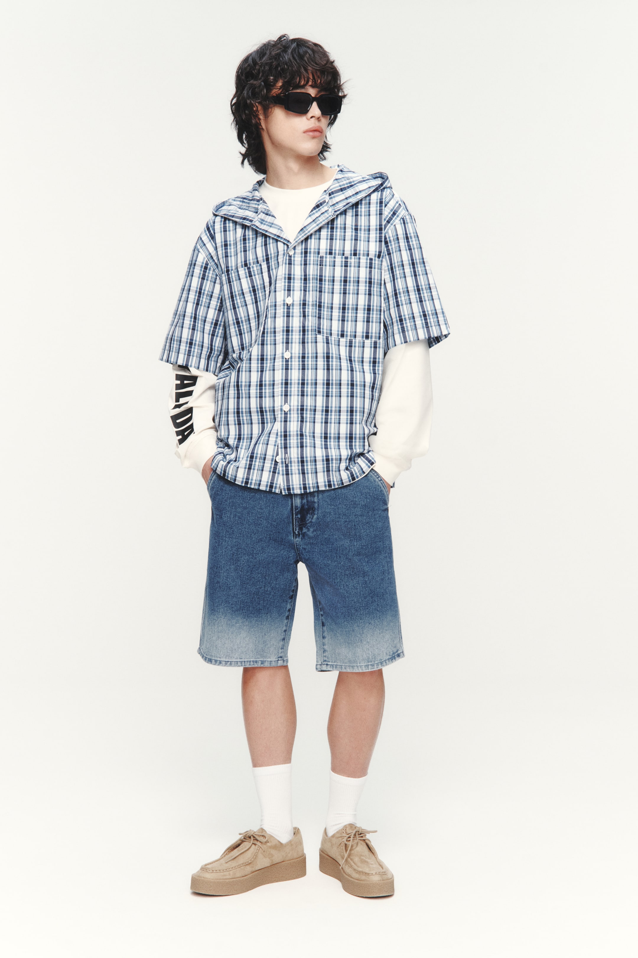 View larger image: The ensemble, showcased from a slightly angled front view, features a blue and white plaid short-sleeve hooded shirt layered over a cream long-sleeve t-shirt with black text on its sleeves. It is paired with faded blue denim shorts, which darken from the waist to the knees, and tan suede-like platform lace-up shoes worn with white crew socks.