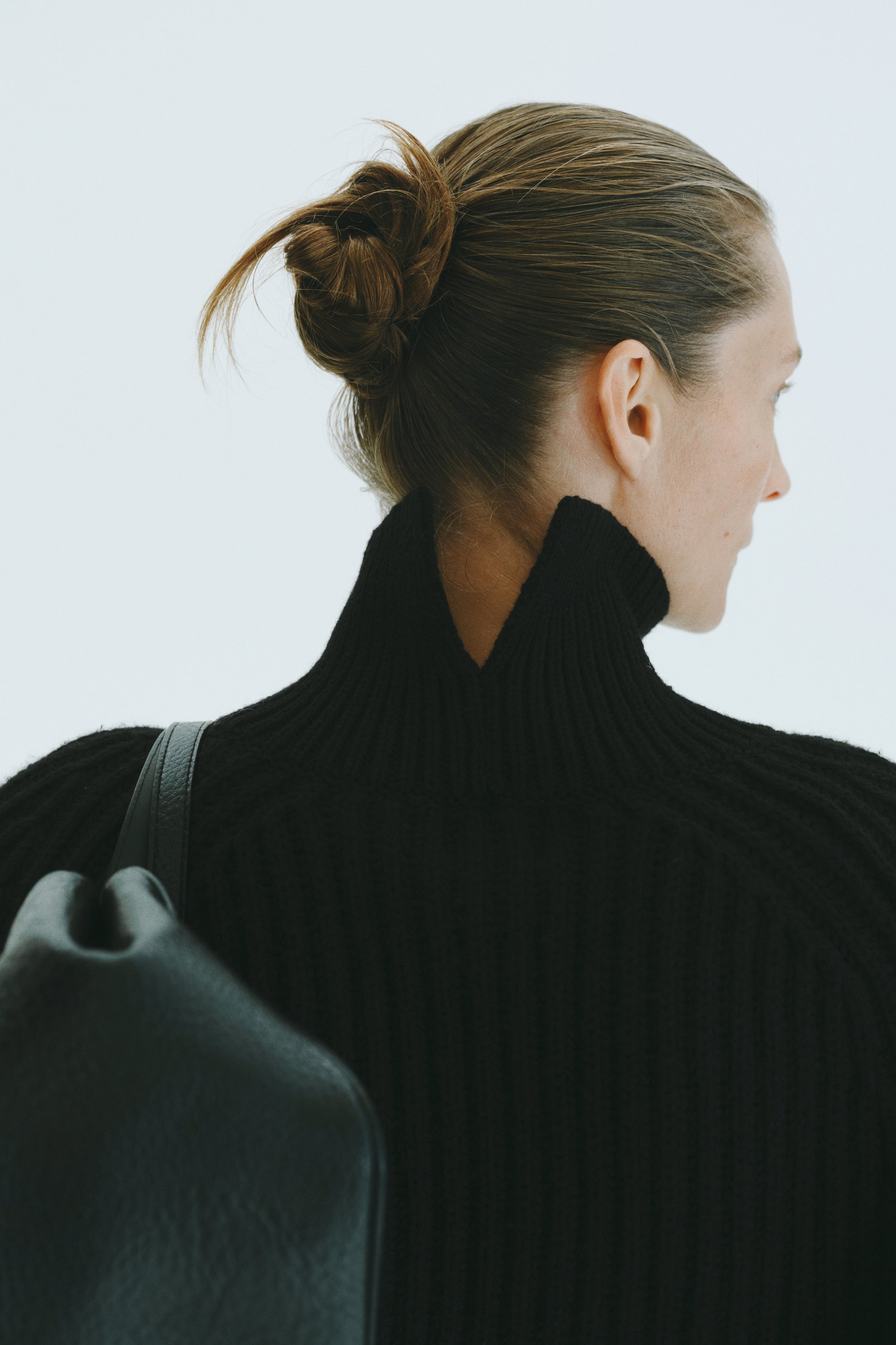 View larger image: The model is wearing a black ribbed knit sweater with a high neck and a V-shaped opening at the back. A black leather bag is worn over the shoulder, with the strap resting on the sweater.