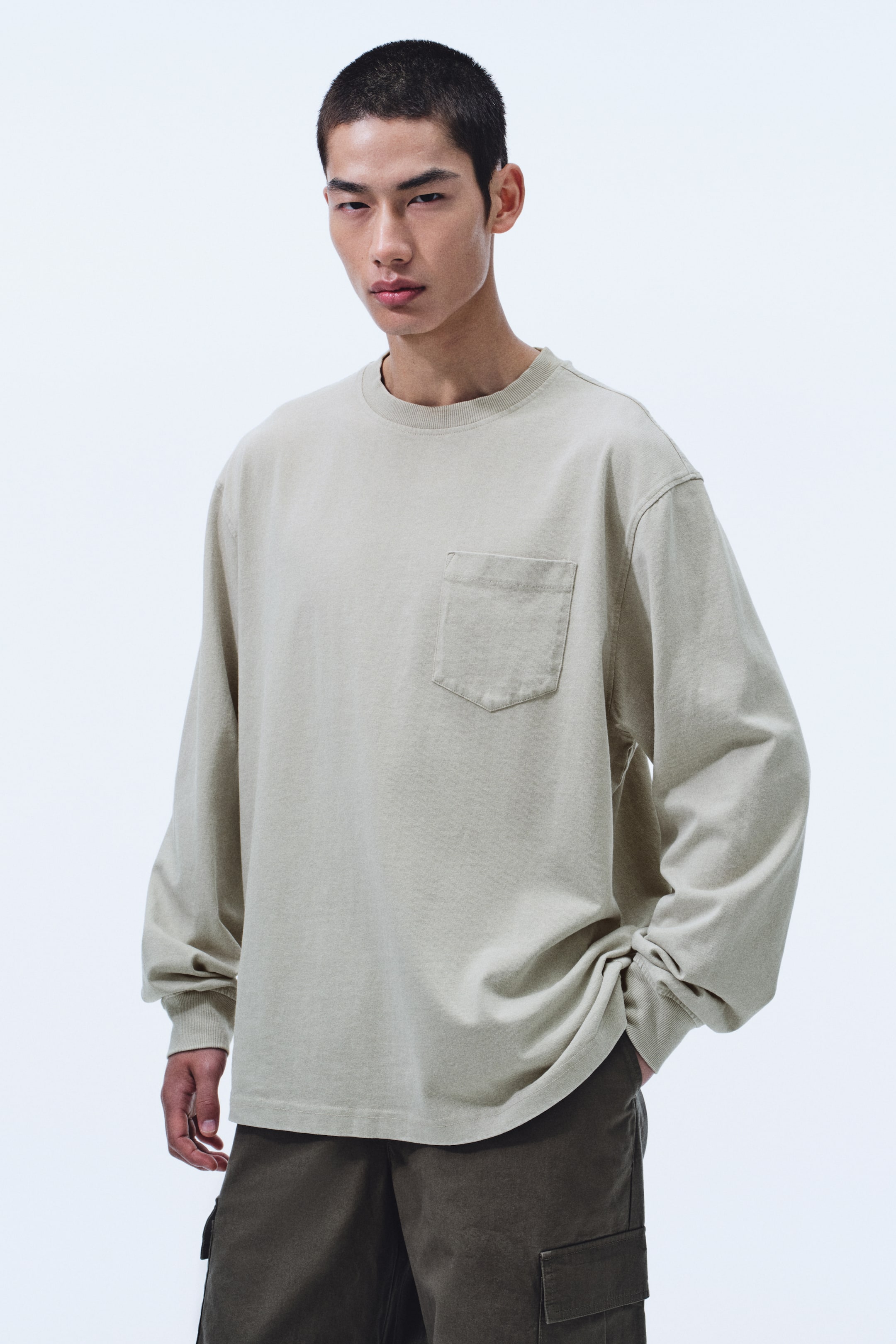 View larger image: A light beige long-sleeved T-shirt is shown from the front at a slight angle, featuring a ribbed crew neck and a single patch pocket on the left chest. It has a relaxed fit with long, loose sleeves and ribbed cuffs, the hem falling over dark pants.