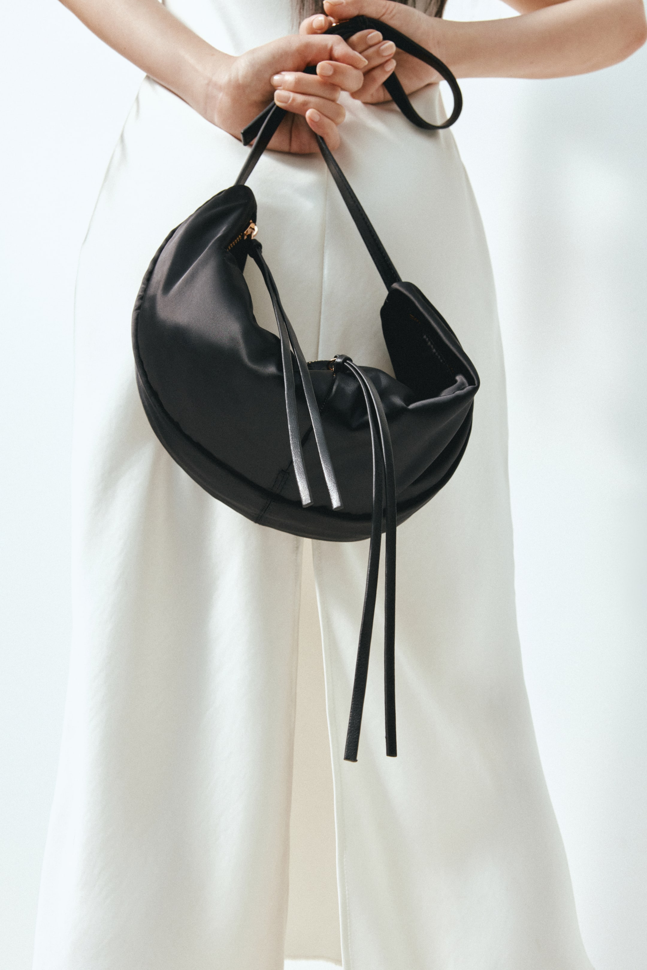 View larger image: A black, crescent-shaped bag with long, dangling zipper pull tabs is being held by the model, who is holding its adjustable strap with both hands. The bag is positioned in front of the model’s white garment.