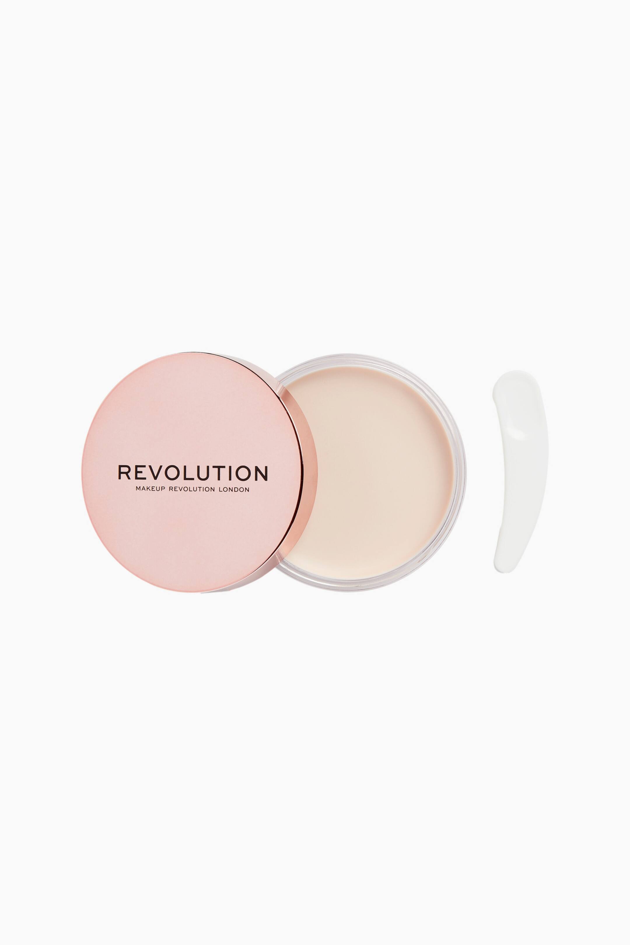 Visa större bild: En öppen rund burk visar en len, ljusbeige balm-primer, med det metalliska roséguldfärgade locket med "REVOLUTION"-logotypen vilande delvis över vänster sida. Ett böjt, slätt vitt krämprov ligger till höger om burken och illustrerar produktens textur.