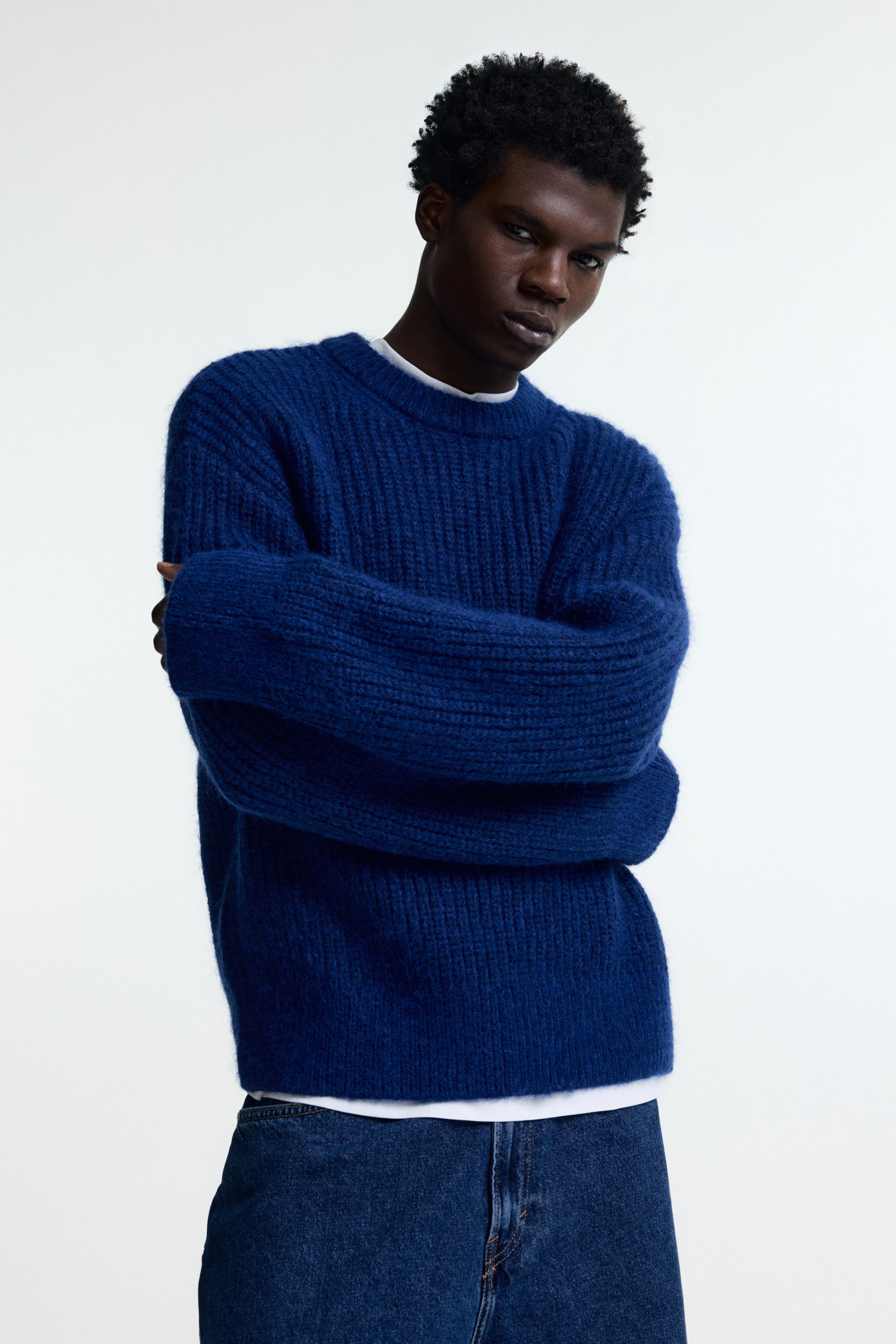 Ampliar la imagen: Un suéter de punto acanalado azul está siendo usado por el modelo, con cuello redondo y mangas holgadas. El modelo cruza los brazos sobre el pecho, causando que las mangas del suéter se doblen, y una camiseta blanca se está usando debajo.