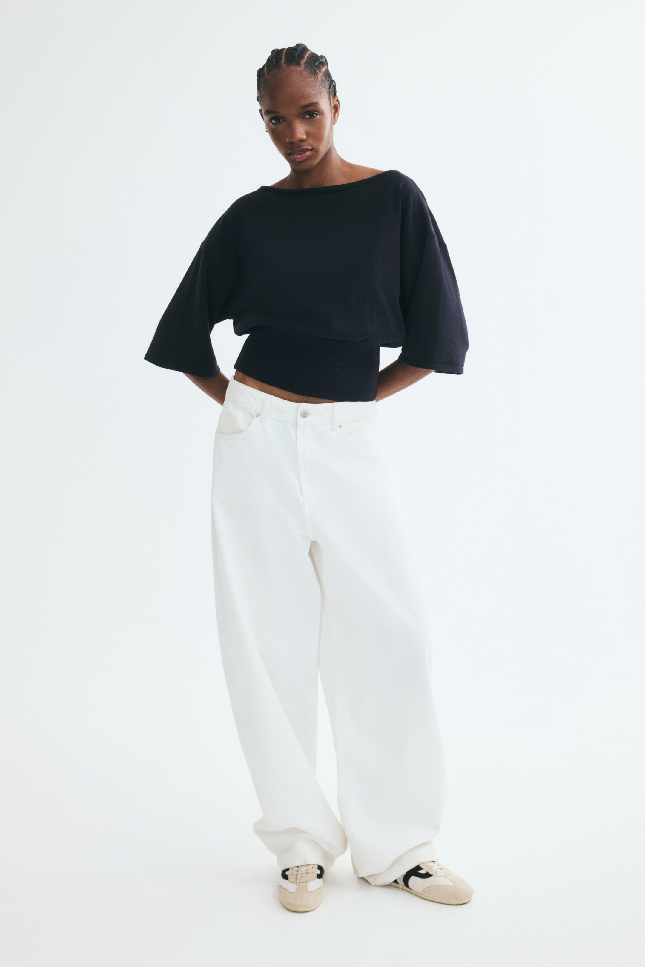 View larger image: A black knit top featuring a boat neck, dropped shoulders, elbow-length sleeves, and a wide, fitted waistband is worn by the model. The top is styled with high-waisted, wide-leg white trousers.