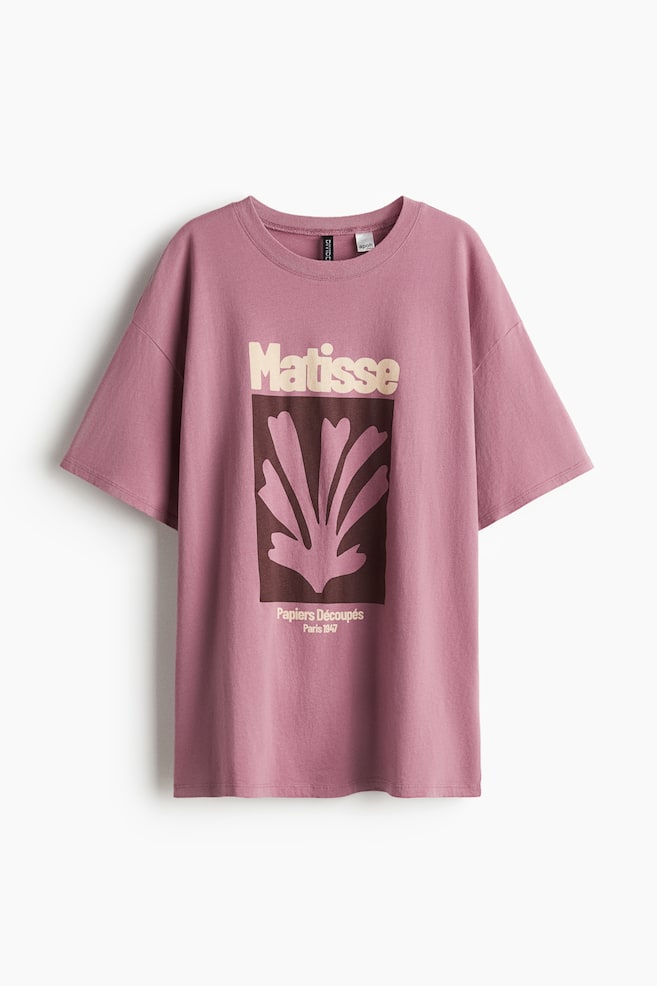 ピンクがかった紫のコットンジャージーのオーバーサイズTシャツは、丸いリブトリムのネックライン、ドロップショルダー、ショートスリーブを特徴とするフロントを見せています。フロントには、中央に茶色の背景、淡いピンクの抽象的な葉の形、そして上に「Matisse」、下に「Papiers Découpés Paris 1947」と書かれた淡いピンクのテキストがある長方形のグラフィックプリントが展示されています。 - 1
