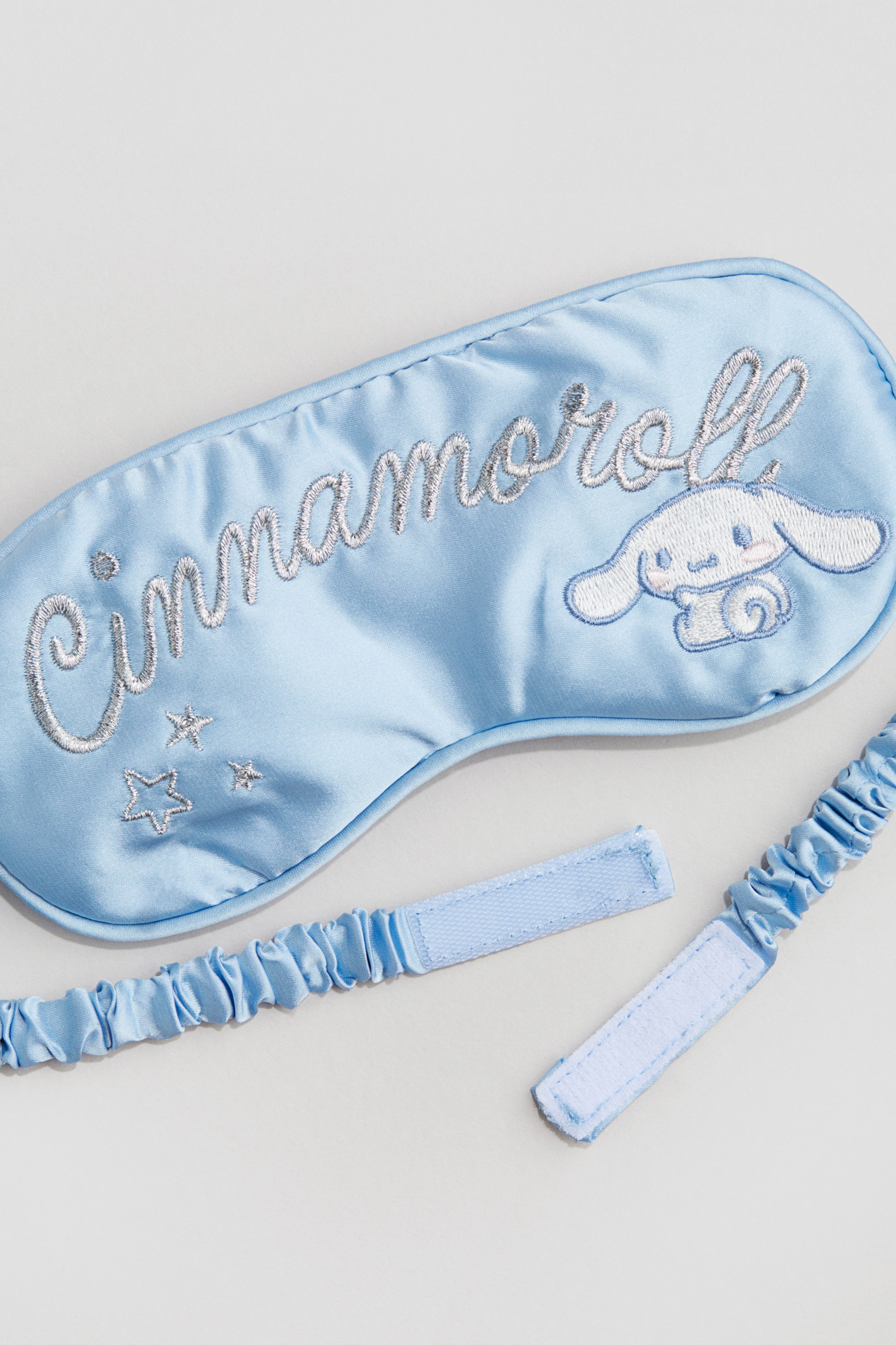 Visualizza immagine più grande: Una maschera per dormire in raso azzurro chiaro è presentata, con 'Cinnamoroll' ricamato in filo grigio argento, accanto a un personaggio bianco e azzurro chiaro e tre stelle grigio argento. Una fascia elastica abbinata azzurro chiaro con chiusure a strappo è posata accanto alla maschera.