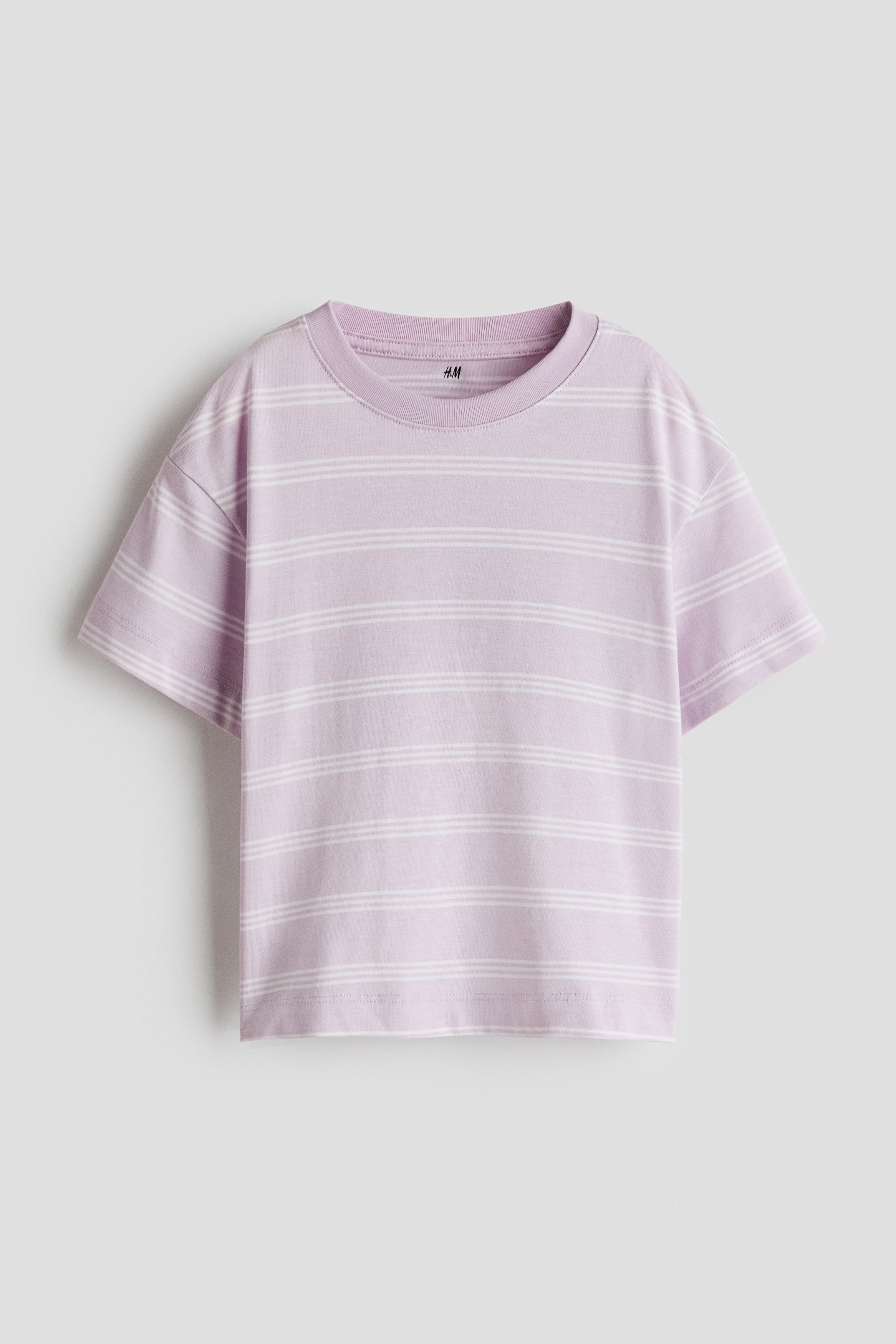The model is wearing a light purple, short-sleeved T-shirt with thin white horizontal stripes, paired with matching solid purple shorts, and is standing with hands resting in the side pockets. The outfit is styled with white crew socks featuring purple stripes and white sneakers with pink accents.