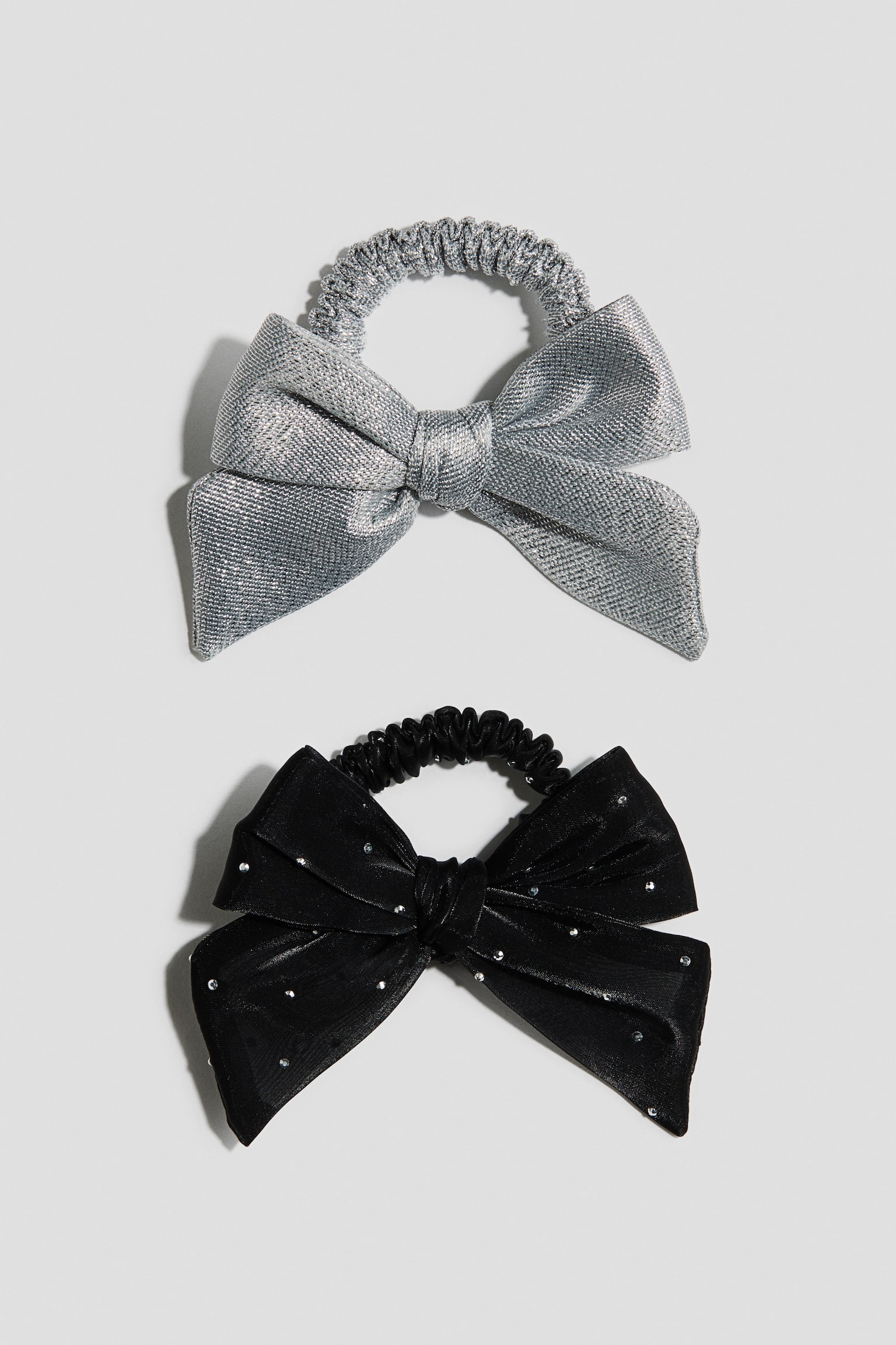 Two elasticated scrunchies featuring decorative bows are shown from the front, with the top scrunchie displaying a textured silver-toned metallic weave and the bottom scrunchie featuring a black fabric embellished with small, glittering rhinestones.