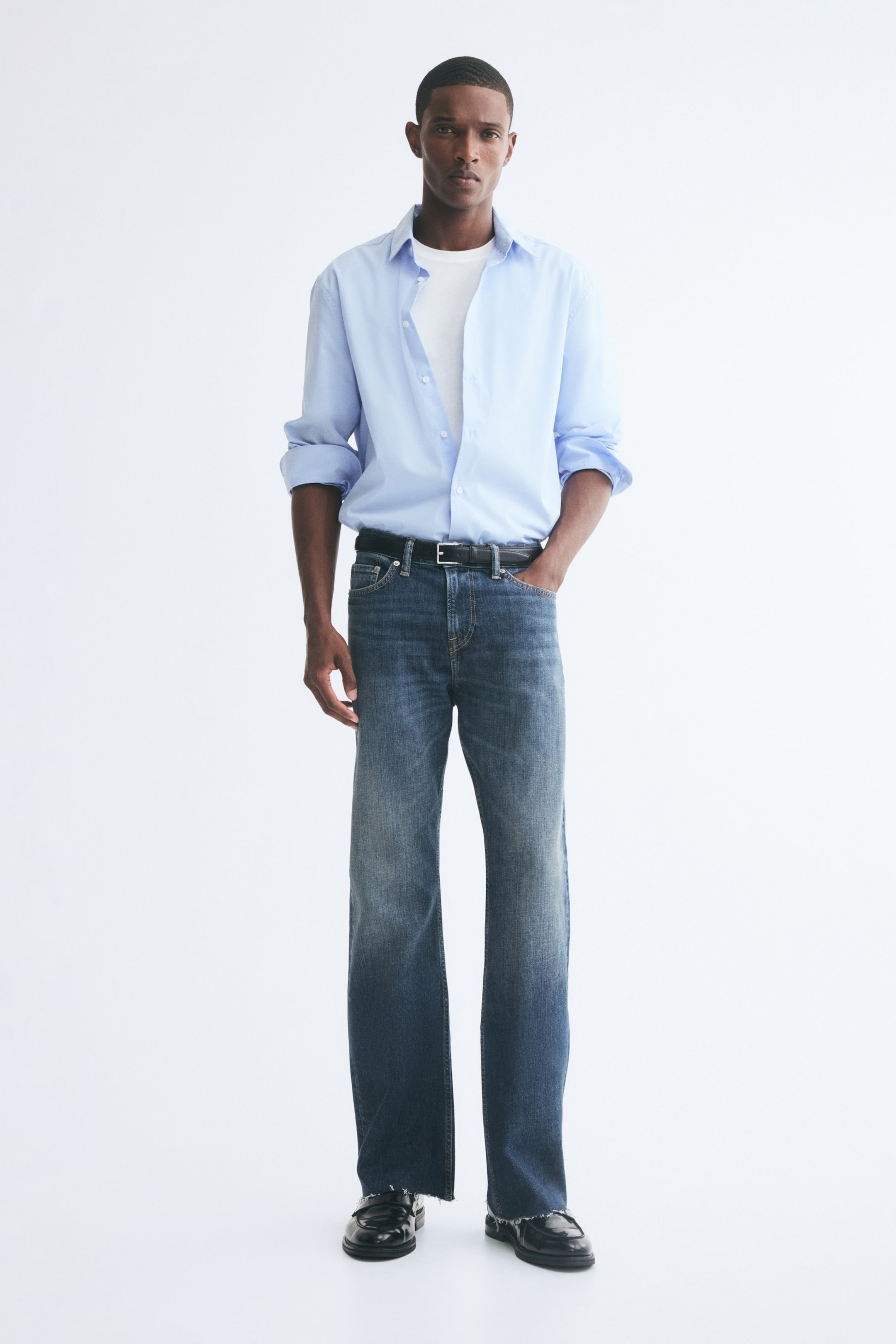 Se større billede: Et par blå denimjeans i medium-vask med lige ben bæres af modellen og har rå kant forneden samt et klassisk design med fem lommer. Jeansene styles med et sort læderbælte, en lyseblå skjorte med knapper lagt over en hvid T-shirt og sorte lædersko.