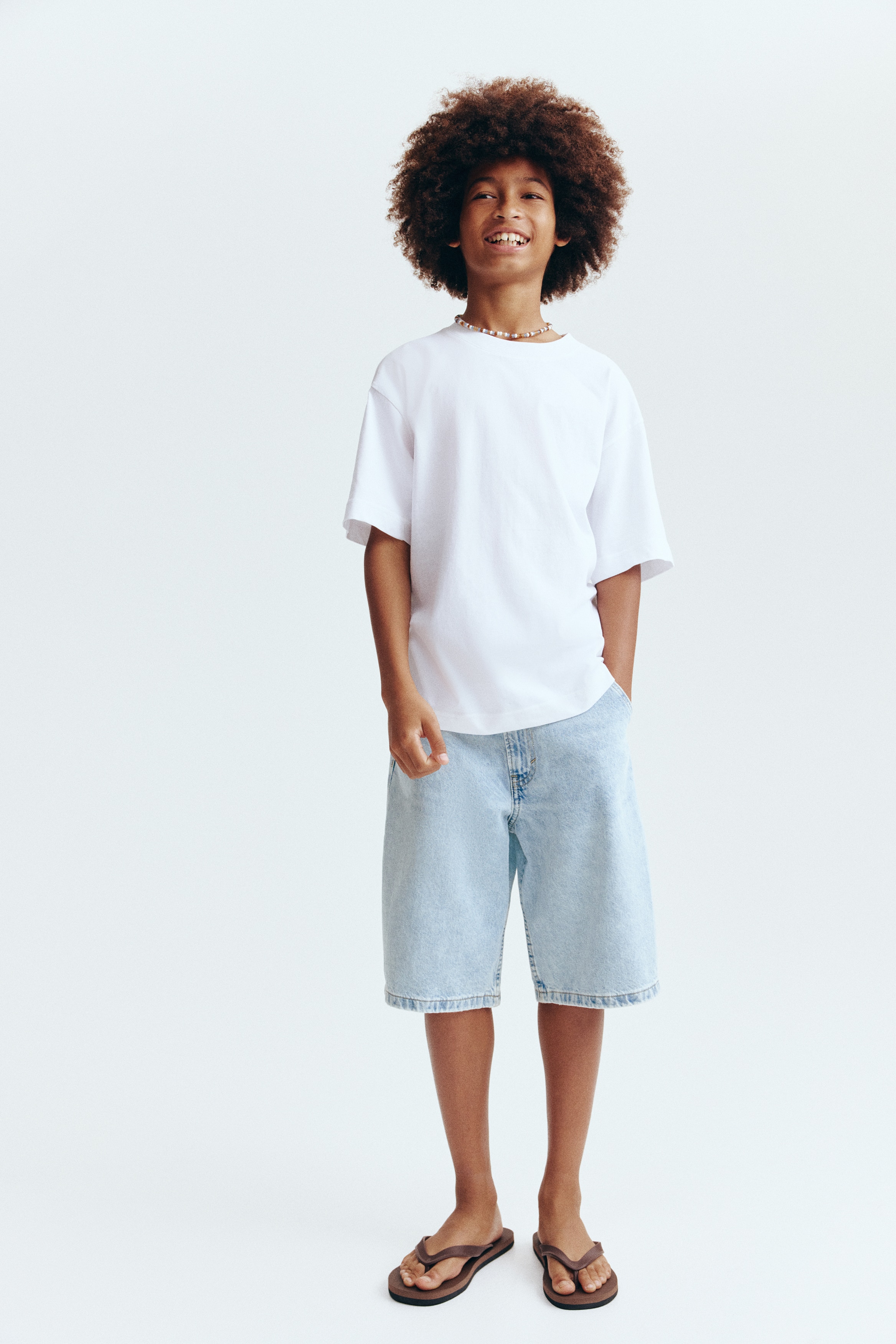 Knee-length shorts are displayed from the front, showcasing a light blue cotton denim with a relaxed fit and an adjustable elasticated waist. They are crafted with a zip fly and button fastening, diagonal front pockets, and a hammer loop on the right side.